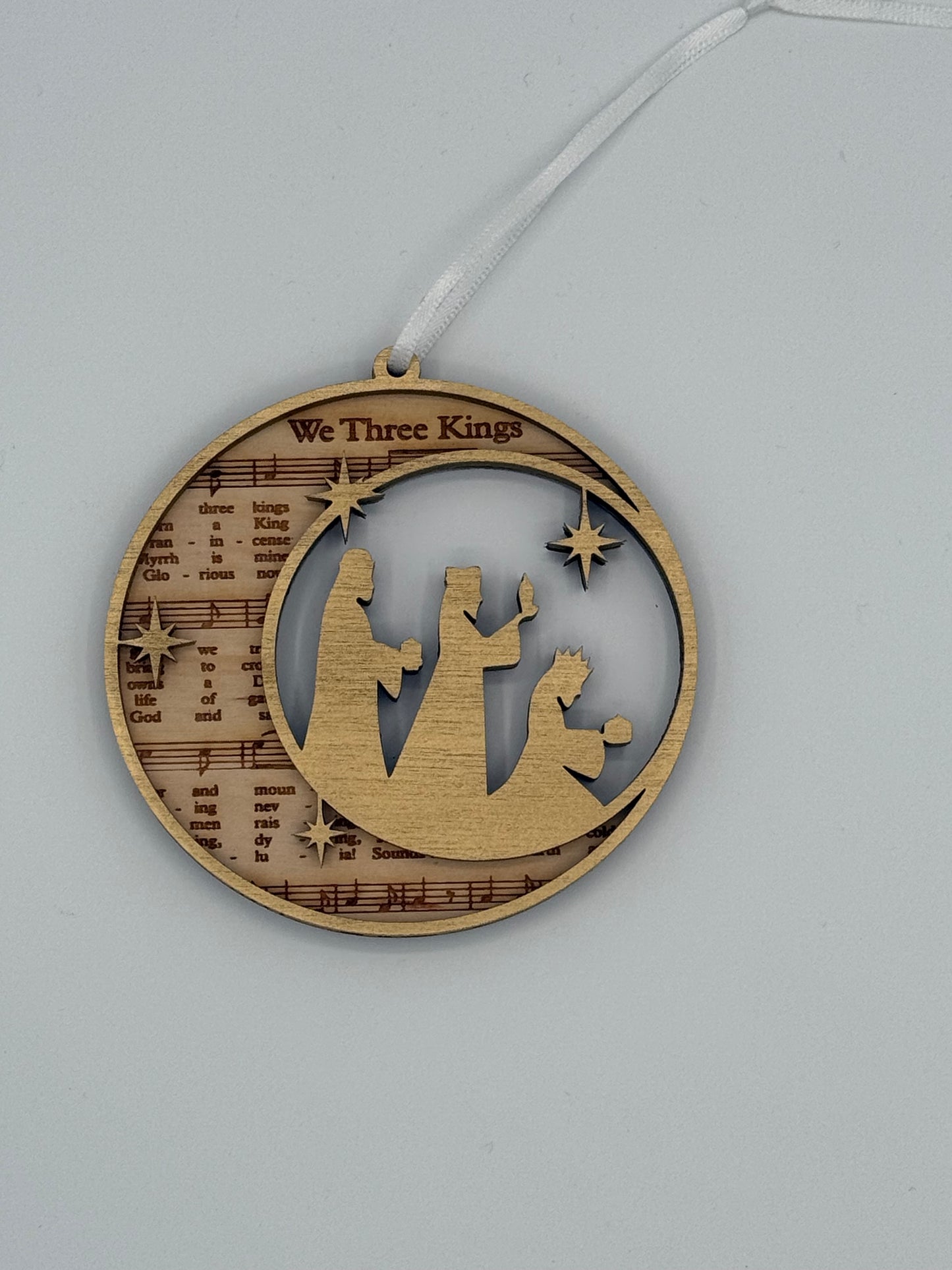 Decorative circular wooden ornament that features 'We Three Kings' bible hymn lyrics engraved in the background with a gold wise men scene overlay.