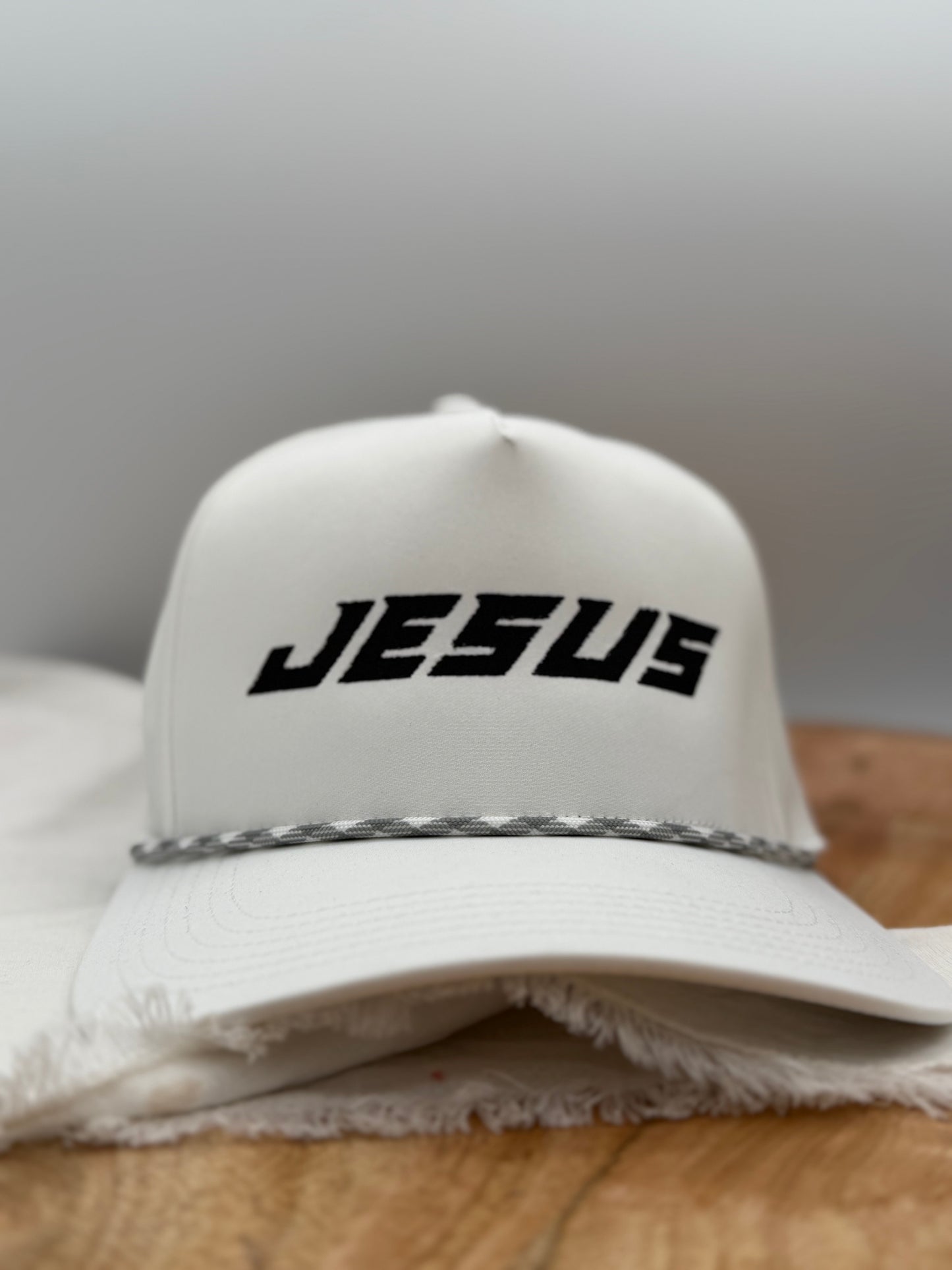 White cap with 'JESUS' embroidered on it in black, featuring a black and white patterned strap, on a wooden surface front facing view of the hat