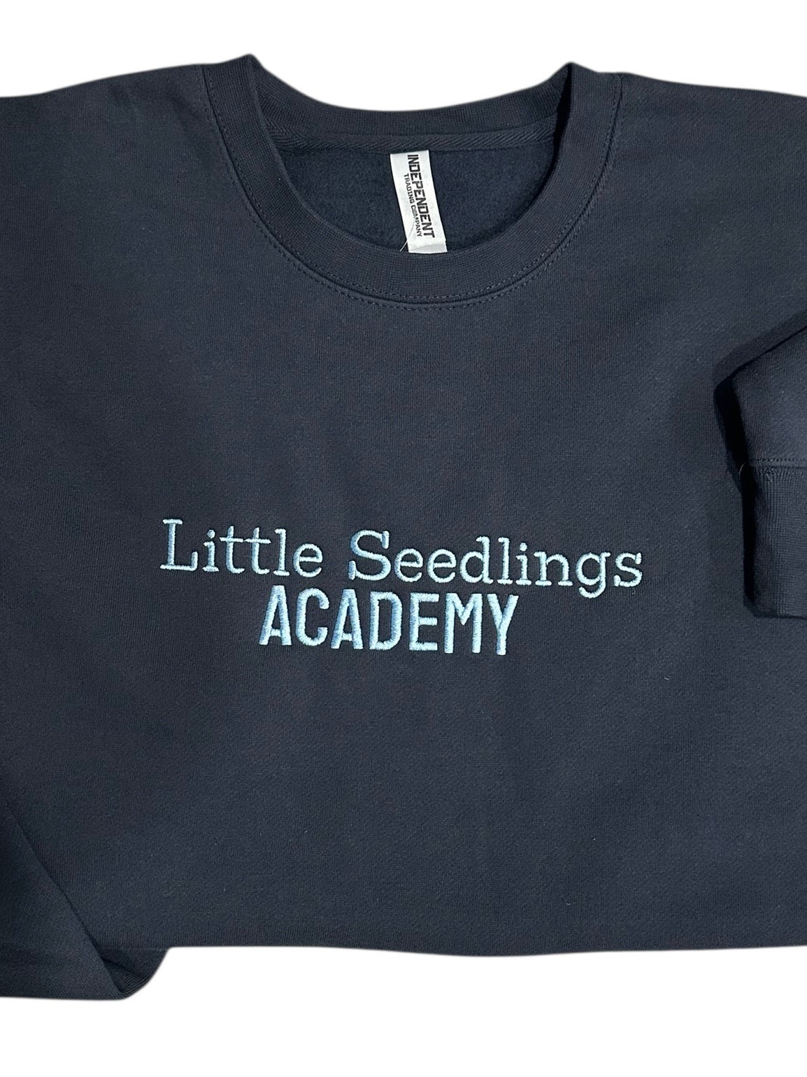 Navy blue sweatshirt with 'Little Seedlings Academy' text embroidered on front center chest in light blue. This is a zoomed in view with focus on the design.