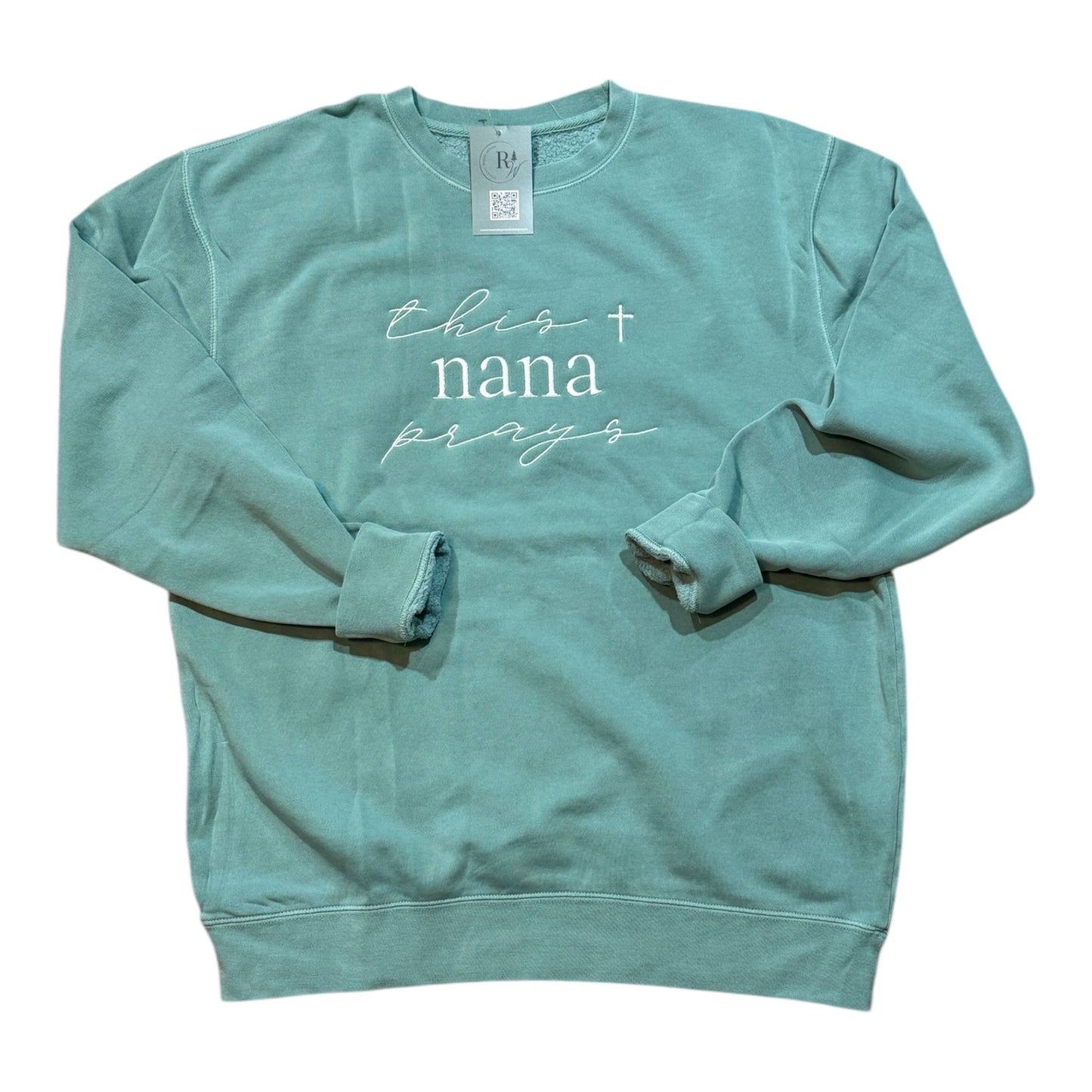 Teal green sweatshirt with the words 'this nana prays' embroidered on the front center chest with a white cross detail on a white background