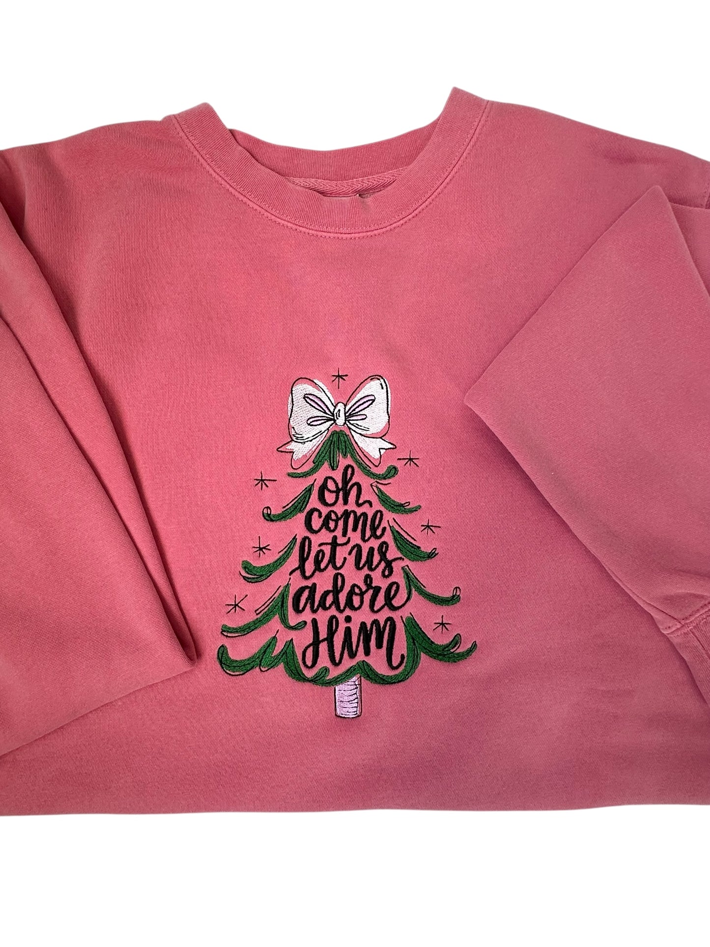 Pink sweatshirt with Christmas tree design and text going through the center of the tree that says 'Oh come let us adore Him' him embroidered front center chest. The tree features a white bow, the tree is green, and the words are black. Sweatshirt is folded in half and sleeves are arranged in a frontal view.
