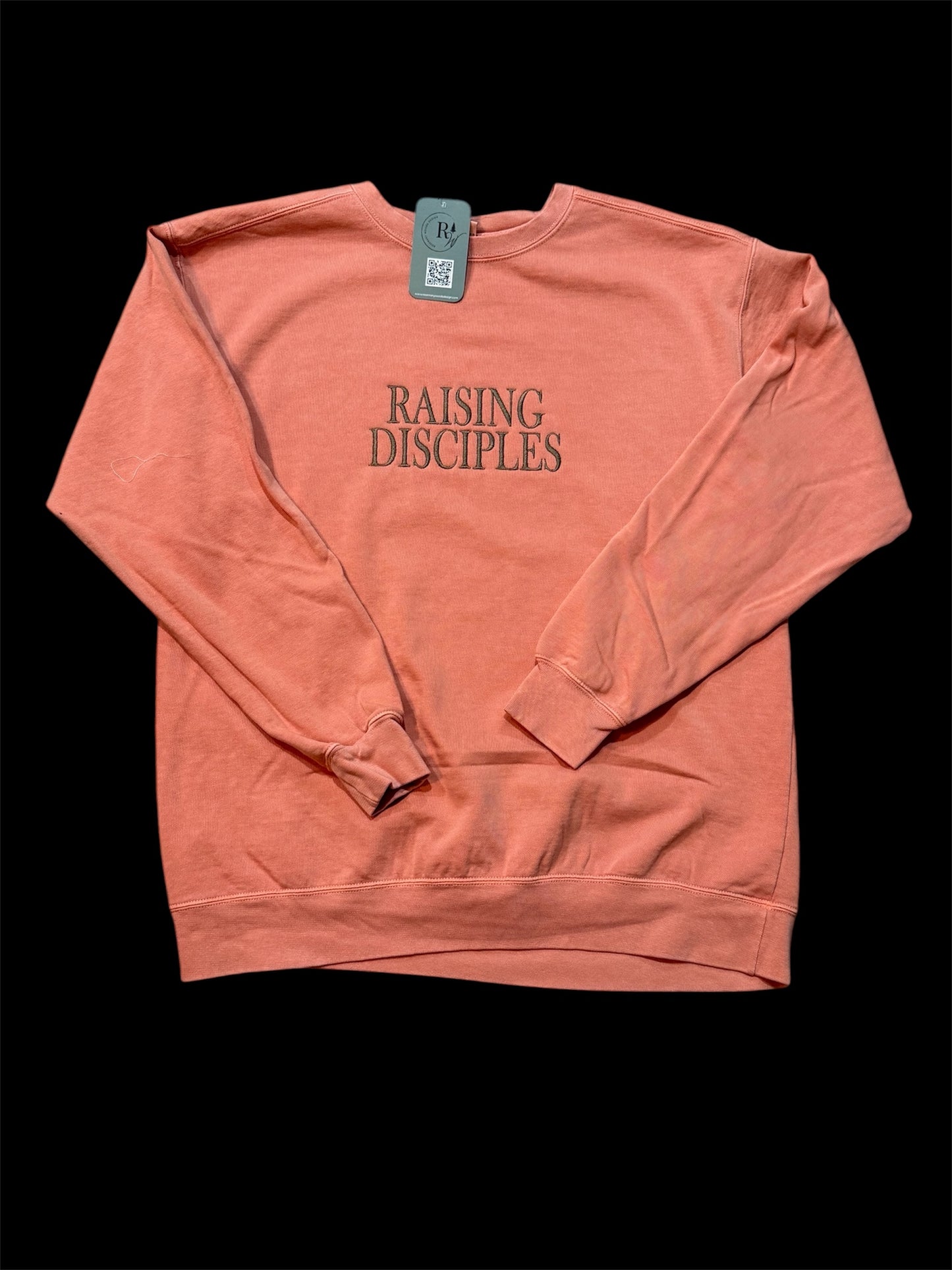 Coral sweatshirt with the text 'Raising Disciples' embroidered in capital letters in brown. This view is zoomed out with the entire sweatshirt showing with sleeves folded on top.