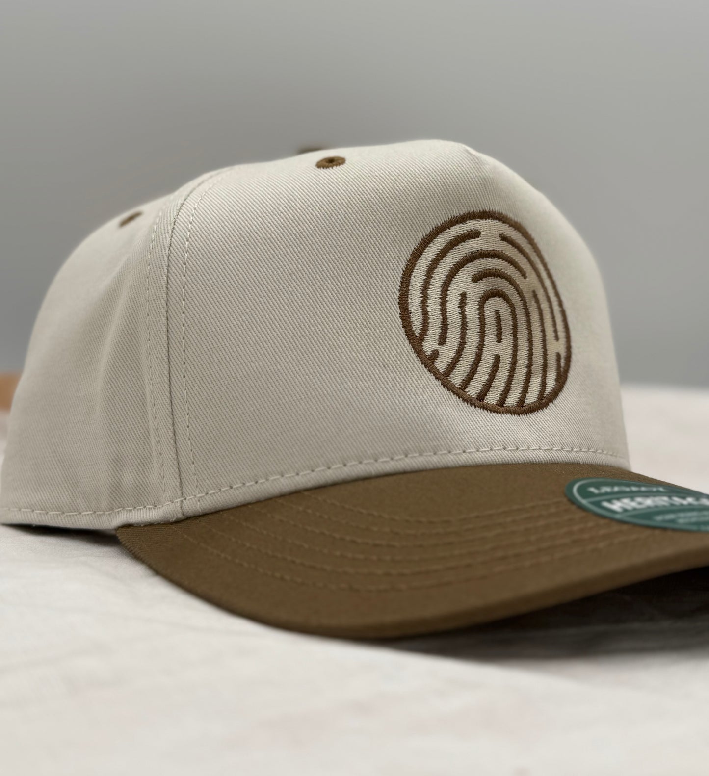 Beige and brown 2 tone hat side profile with a Soma church fingerprint logo embroidered in brown on a gray background