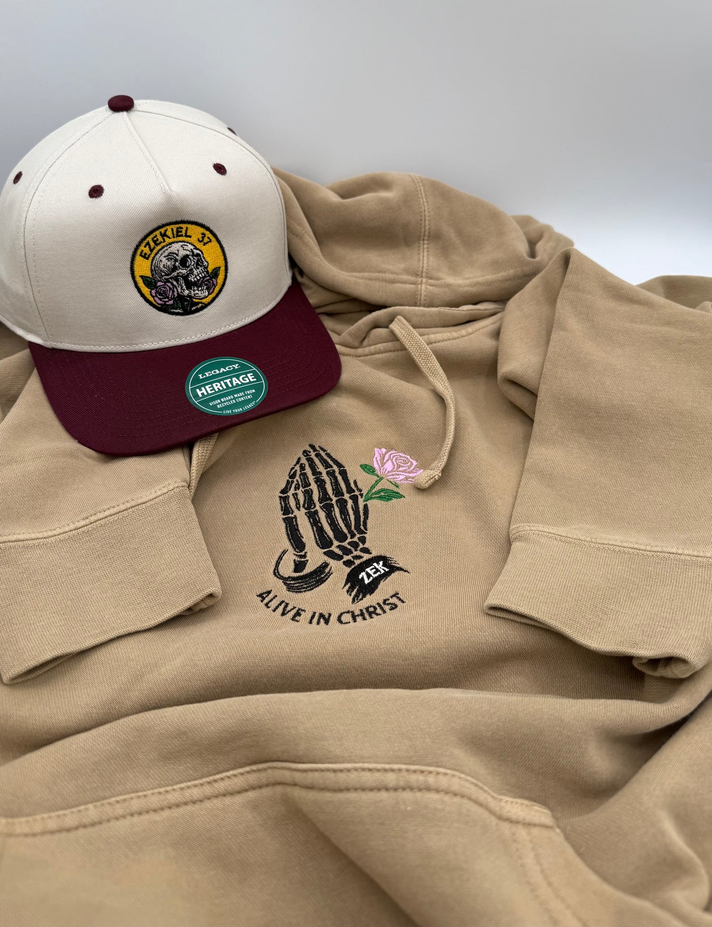 Bundle picture with a tan hooded sweatshirt with skeleton hands holding a rose embroidered in black and pink on the center chest. The words 'Alive in Christ' are embroidered underneath. A tan and maroon two toned hat with a skull holding a rose is embroidered in a circular pattern with the words "Ezekiel 37".