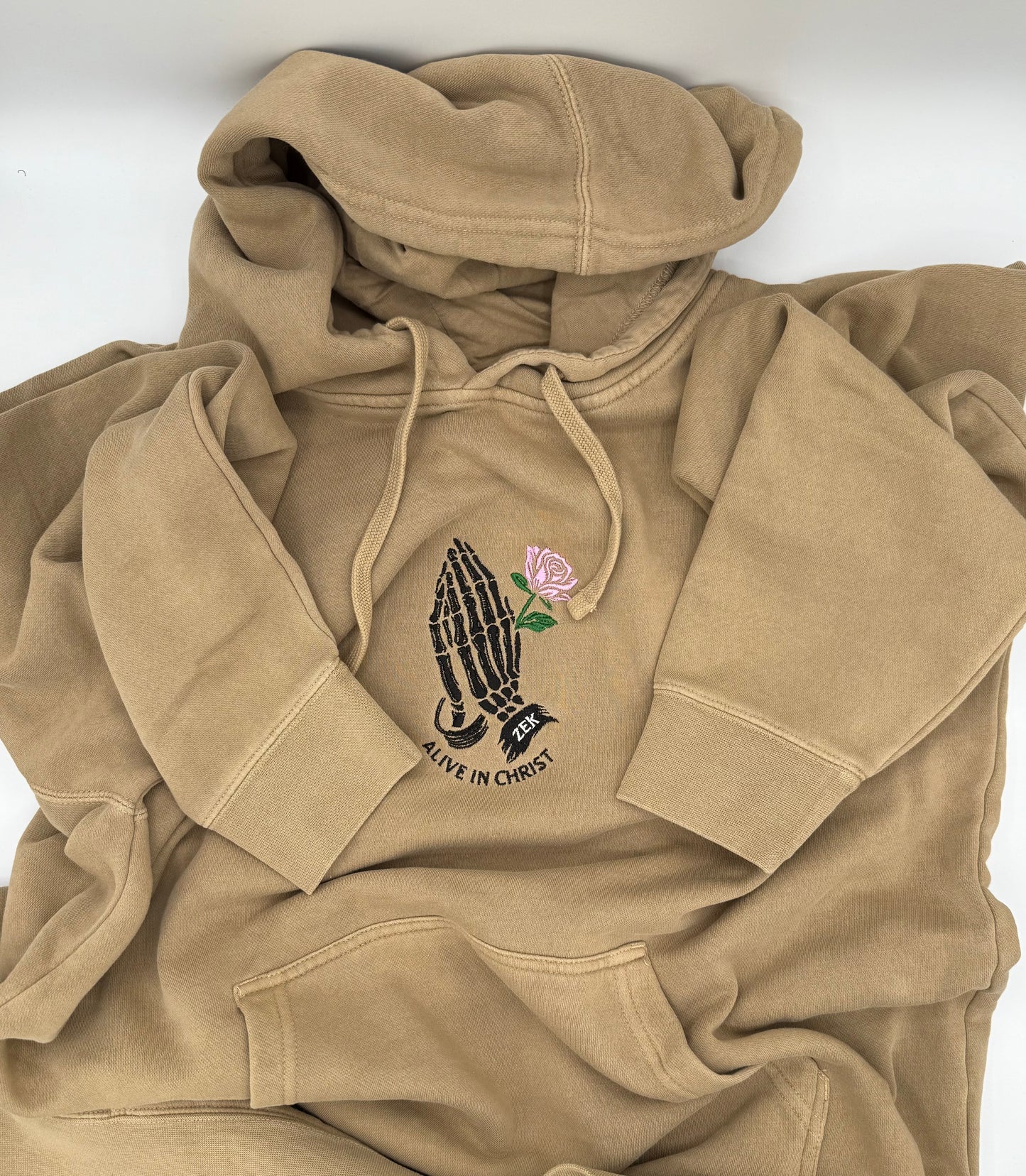 Tan hooded sweatshirt with skeleton hands holding a rose embroidered in black and pink on the center chest. The words 'Alive in Christ' are embroidered underneath.