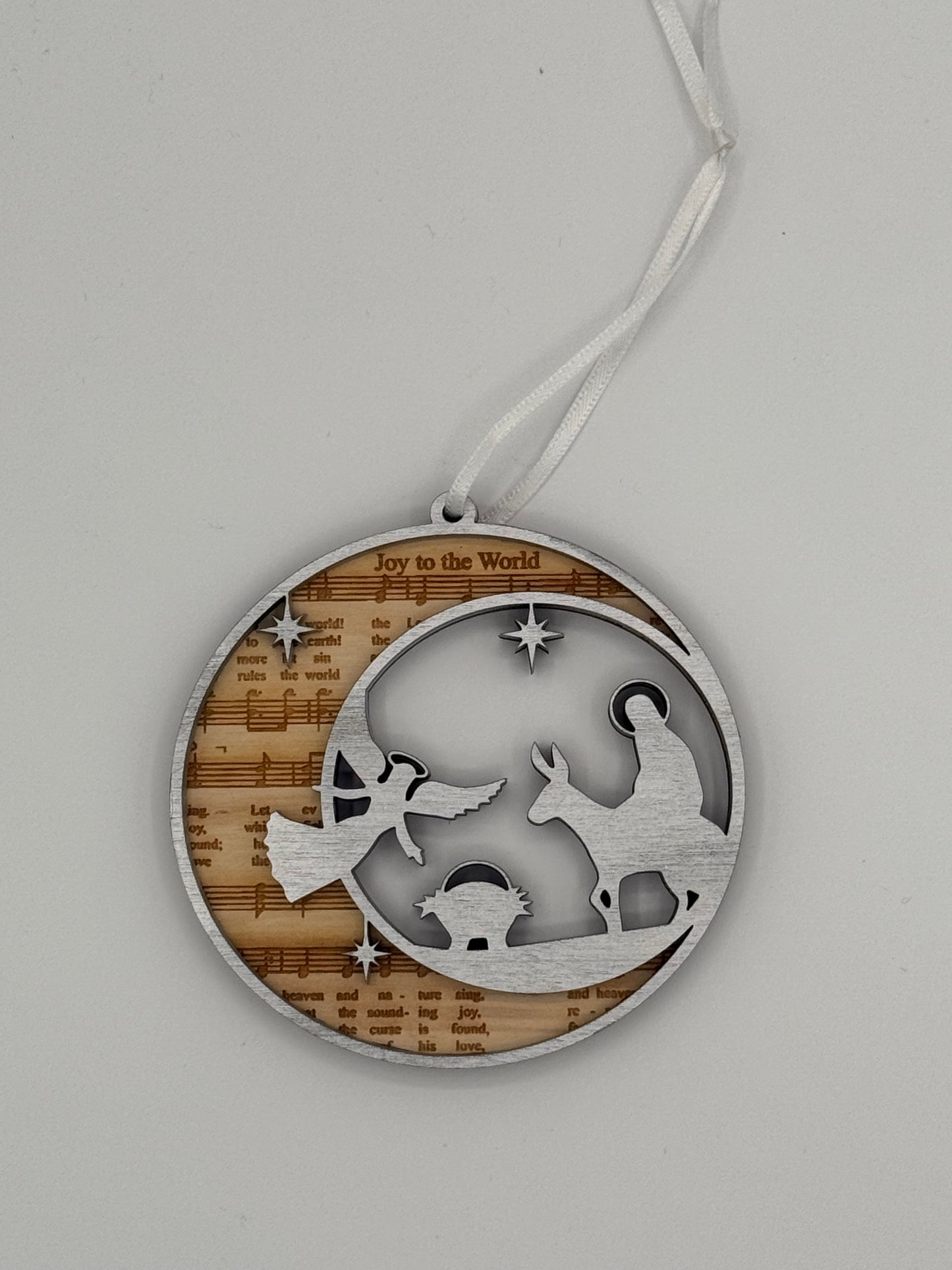 Decorative circular wooden ornament that features 'Joy to the Word' bible hymn lyrics engraved in the background with a silver angel and manger scene overlay.