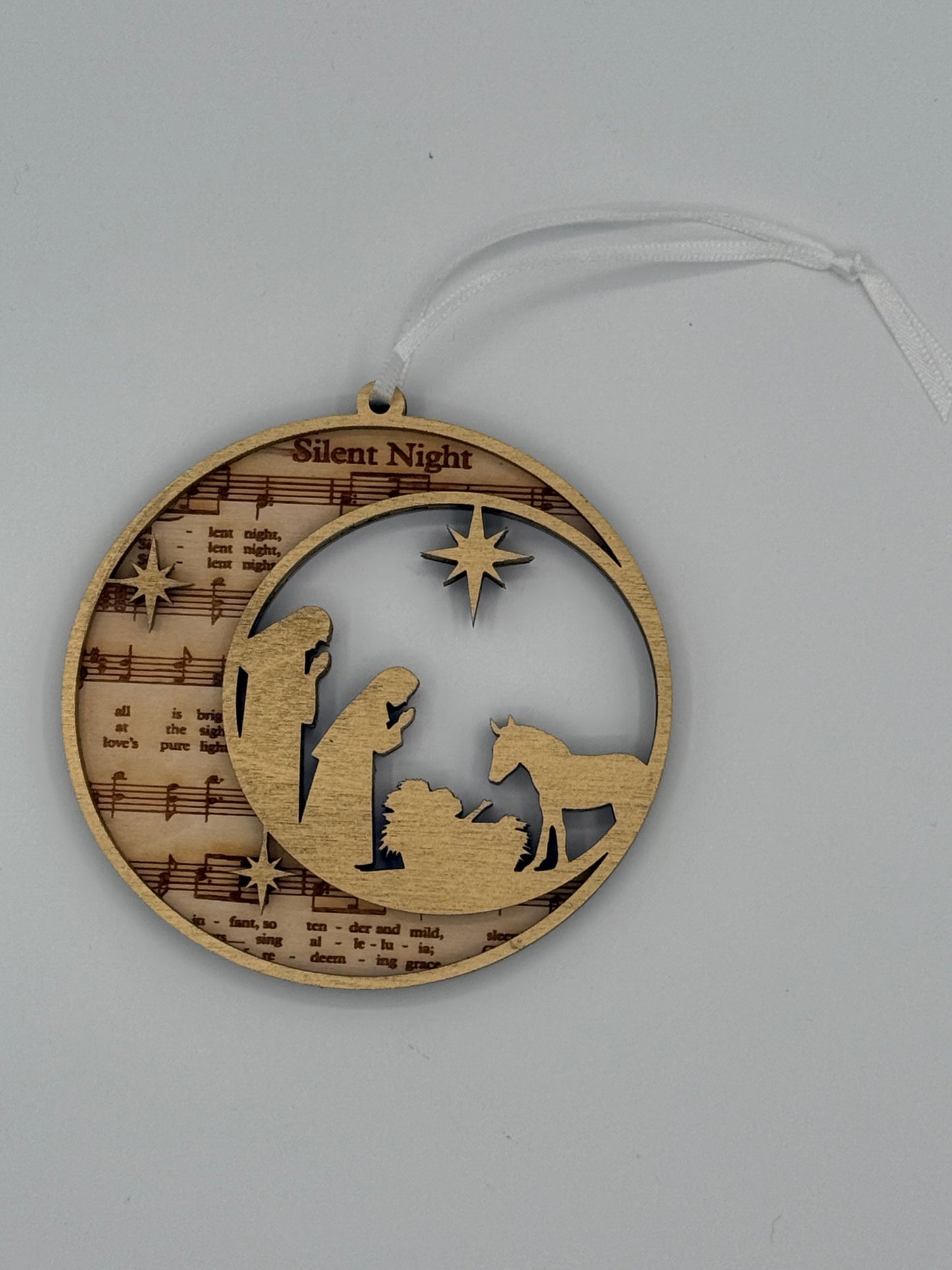 Decorative circular wooden ornament that features 'Silent Night' bible hymn lyrics engraved in the background with a gold manger scene overlay.