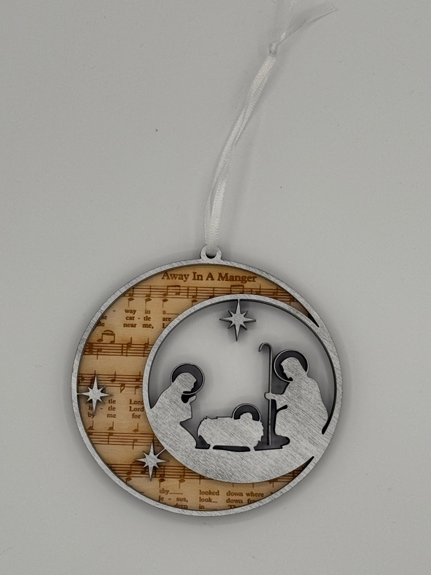 Decorative circular wooden ornament that features 'Away in a Manager' bible hymn lyrics engraved in the background with a silver manger scene overlay.