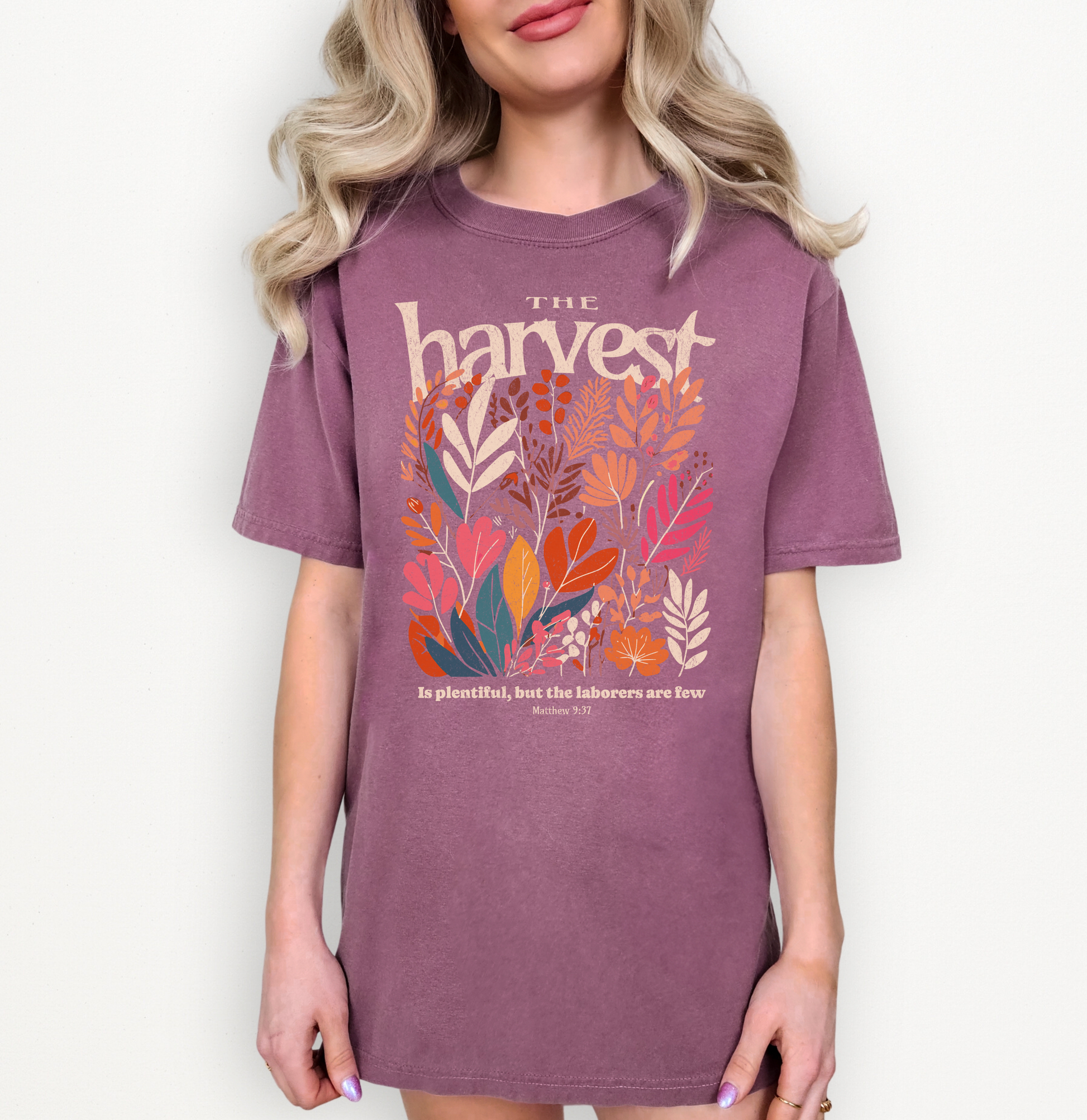 A woman wearing a mauve-purple graphic T-shirt with the word 'harvest' in cream and a colorful arrangement of autumn leaves in orange, red, teal, and cream. Smaller text below reads 'is glorious, but the laborers are free' from Exodus 23:6.