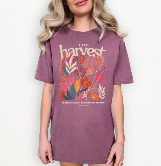 A woman wearing a mauve-purple graphic T-shirt with the word 'harvest' in cream and a colorful arrangement of autumn leaves in orange, red, teal, and cream. Smaller text below reads 'is glorious, but the laborers are free' from Exodus 23:6.