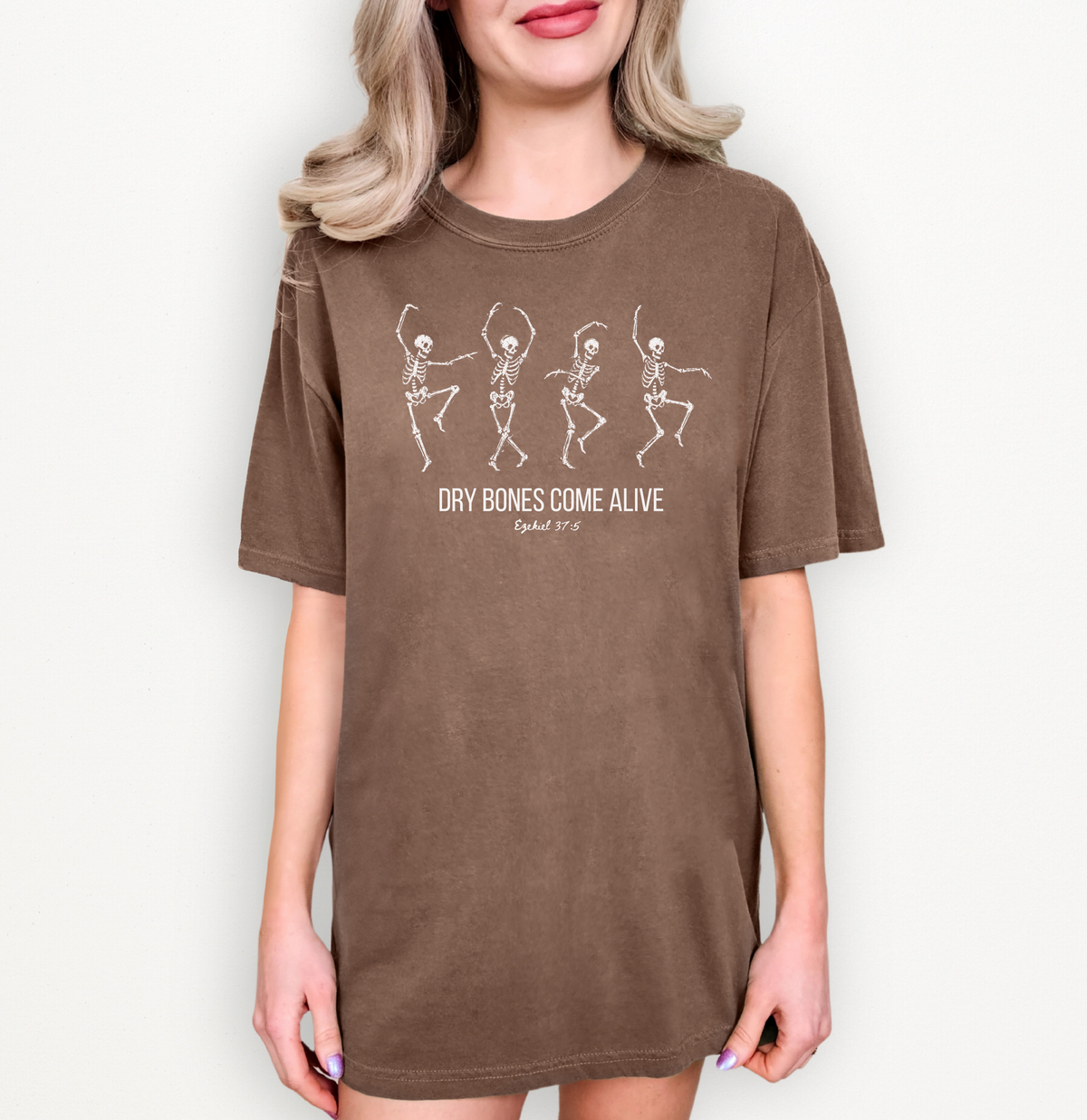 A woman wearing an oversized brown t-shirt dress with a white graphic of dancing skeletons and the text 'DRY BONES COME ALIVE' and 'Ephesians 3:14' on a light off-white background.