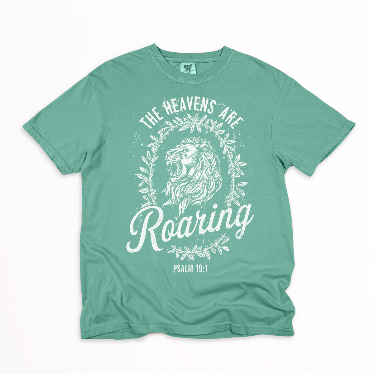 A mint green crewneck t-shirt with a white graphic print featuring a roaring lion, floral wreath, and the text 'THE HEAVENS ARE ROARING' and 'PSALM 15:1'.