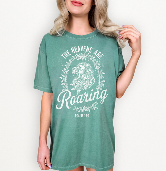 A woman wearing an oversized green graphic T-shirt with a white lion and text design reading 'The Heavens Are Roaring' and 'PSALM 19:1'.