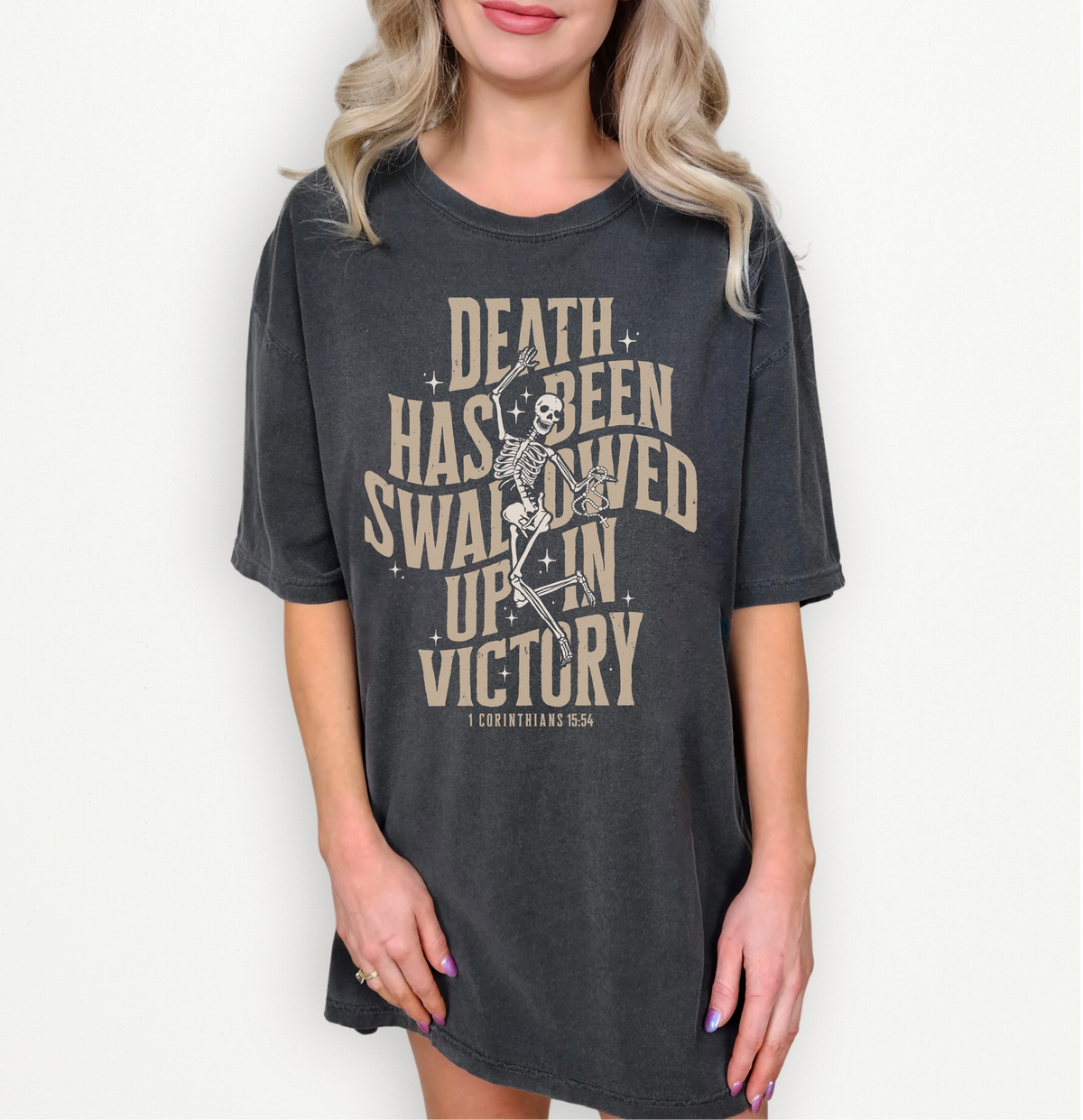 A woman wearing an oversized dark charcoal gray graphic t-shirt with a vintage beige print of a skeleton and the text 'DEATH HAS BEEN SWALLOWED UP IN VICTORY' and 'COUNTRY USA'.