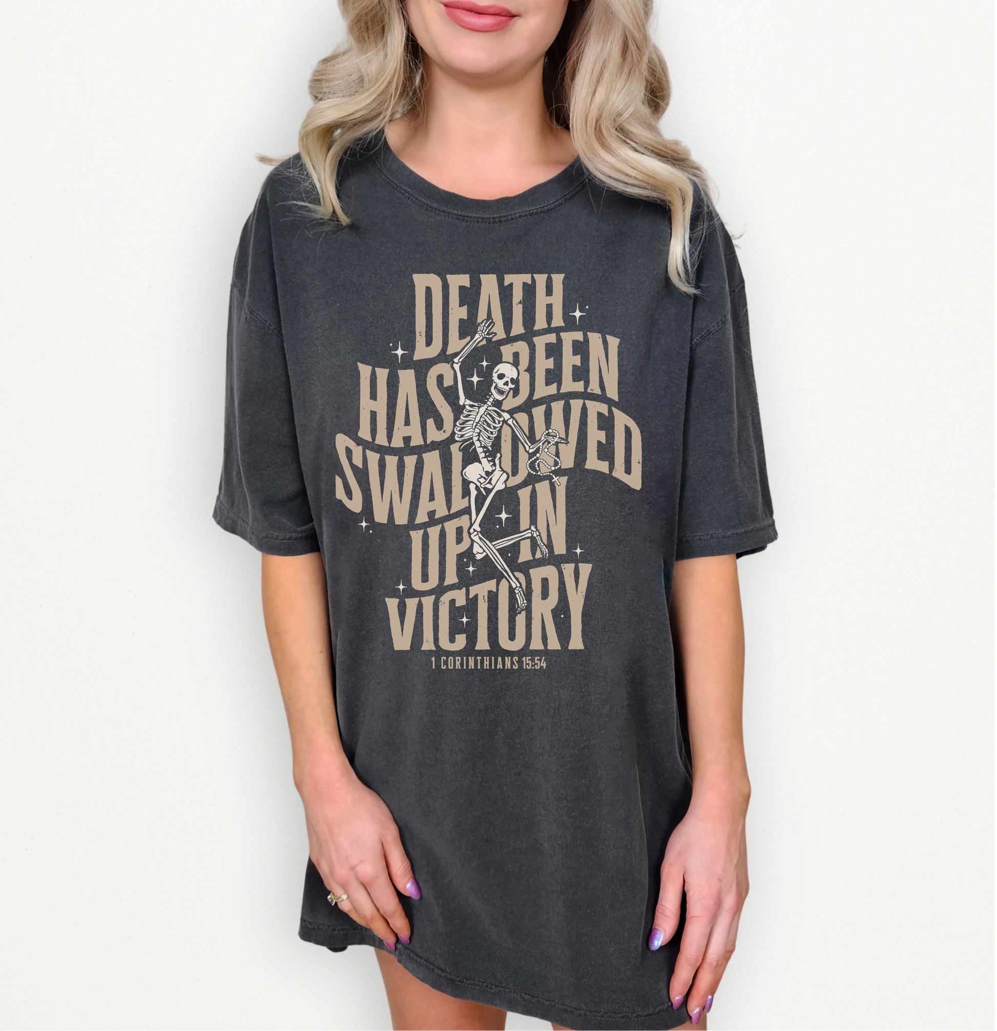 A woman wearing an oversized dark charcoal gray graphic t-shirt with a vintage beige print of a skeleton and the text 'DEATH HAS BEEN SWALLOWED UP IN VICTORY' and 'COUNTRY USA'.