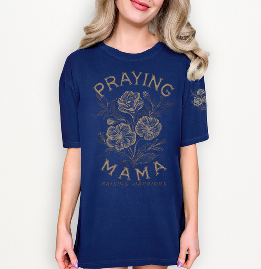 A woman wearing a navy blue short-sleeve t-shirt with a distressed gold graphic that reads 'PRAYING MAMA' and 'RAISING WARRIORS' around a floral illustration on the front.