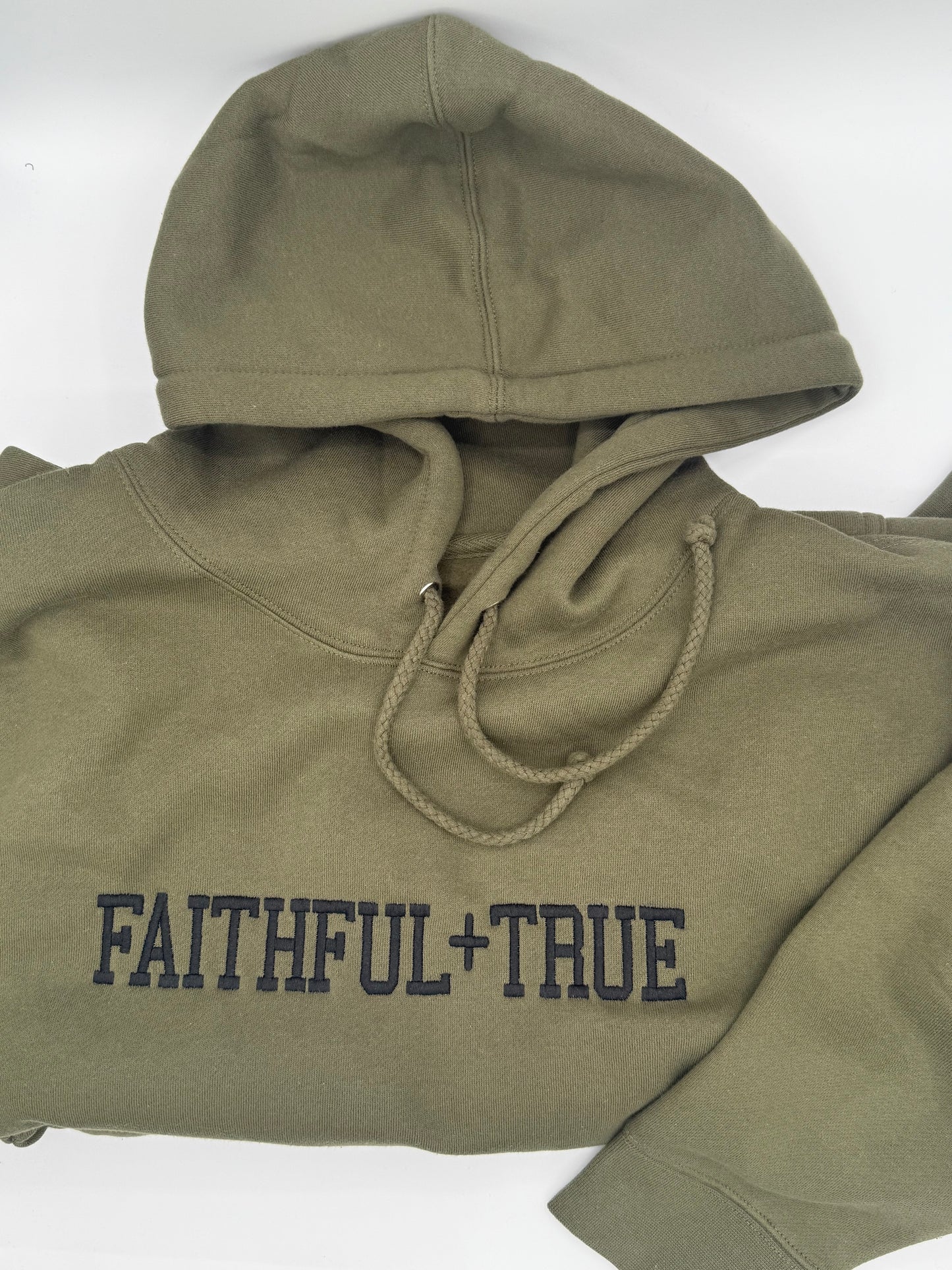 A tan hooded sweatshirt with the words 'Faithful + True' embroidered in black on the front center chest. This is a zoomed in view of the embroidered design with sleeve folded for display.