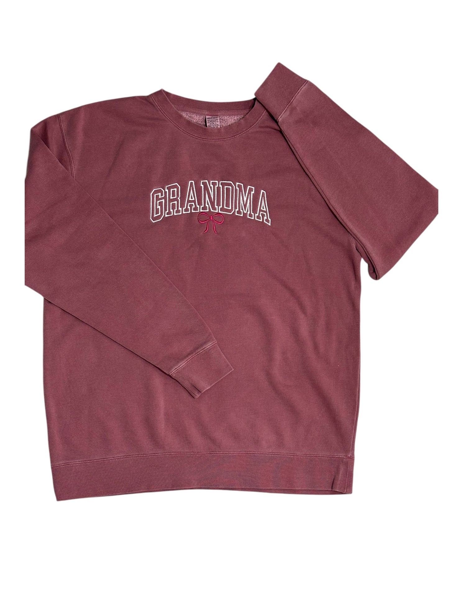 Maroon sweatshirt with the text 'Grandma' embroidered in a white varsity outline font. This design features a maroon embroidered bow underneath. This view is zoomed out with the entire sweatshirt showing and the sleeves folded in opposite directions on top.