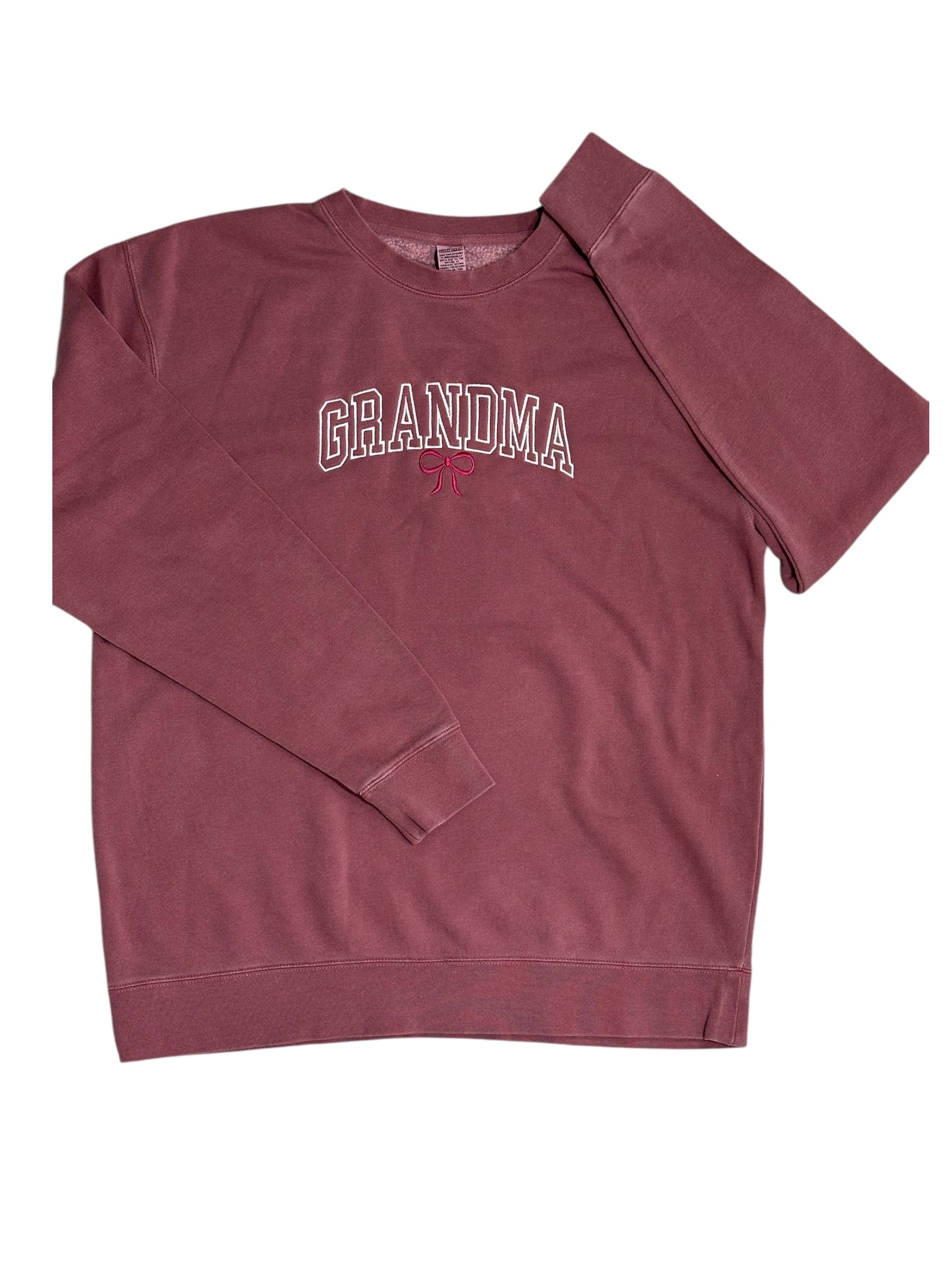Maroon sweatshirt with the text 'Grandma' embroidered in a white varsity outline font. This design features a maroon embroidered bow underneath. This view is zoomed out with the entire sweatshirt showing and the sleeves folded in opposite directions on top.