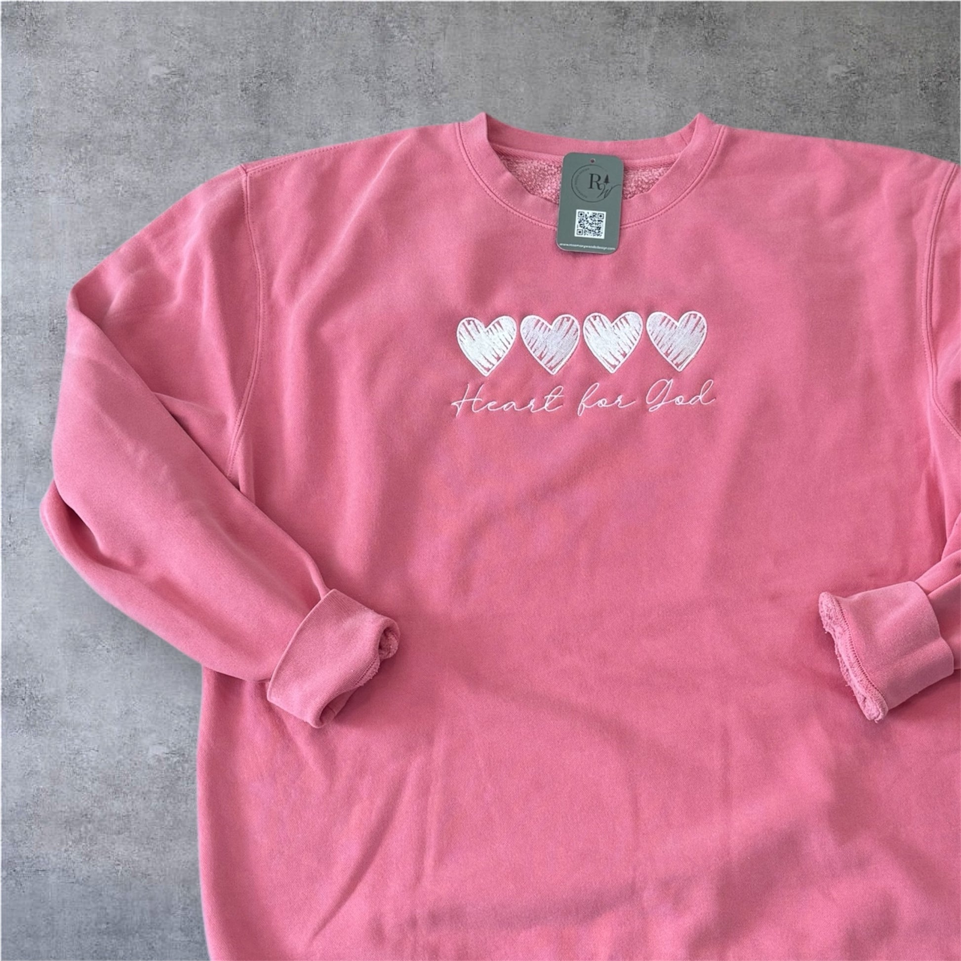Pink long-sleeve sweatshirt with heart designs and text that says Heart for God on a gray surface more zoomed in view of design