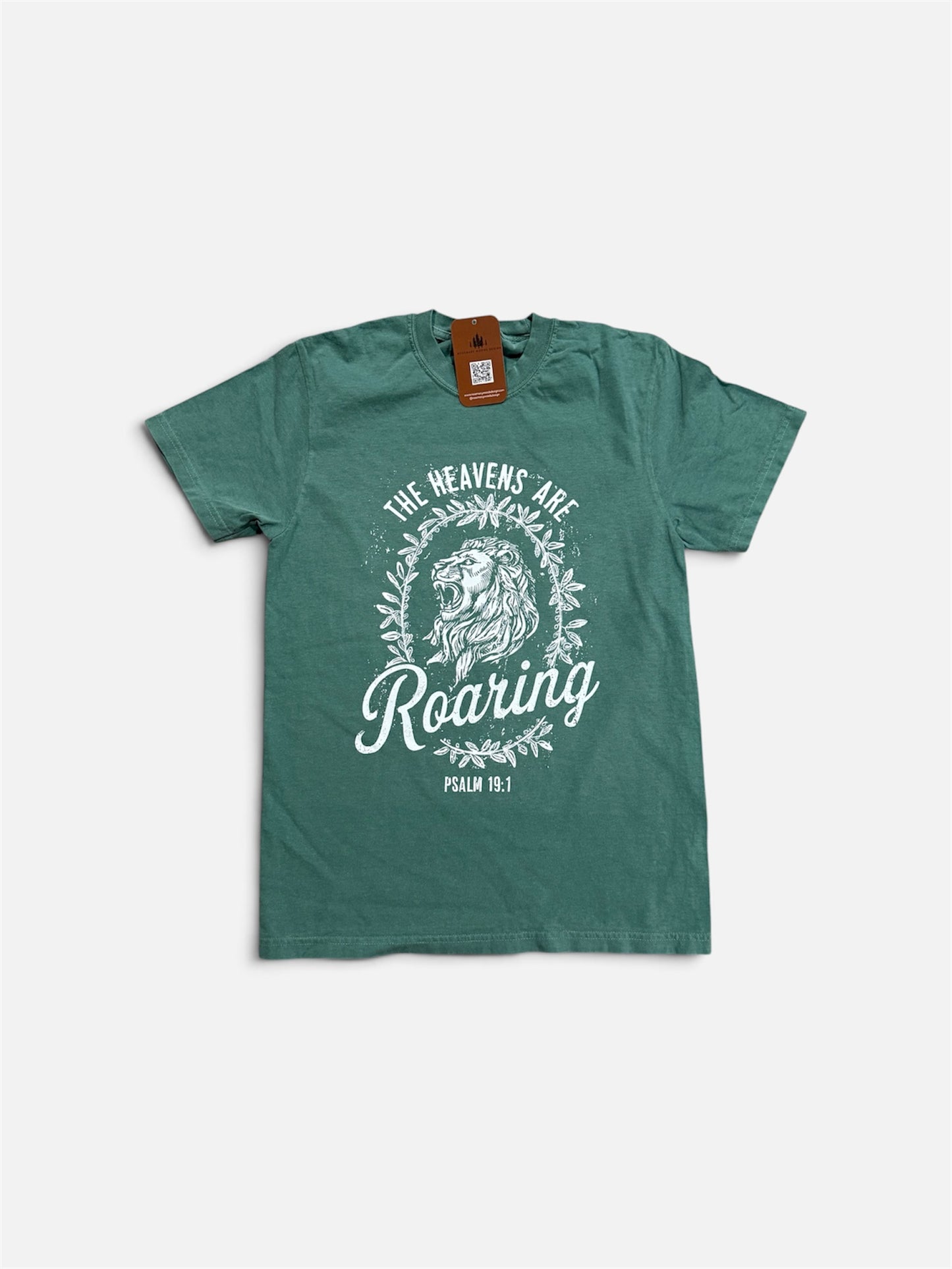 A green short-sleeve t-shirt with a white graphic print featuring a lion, a wreath, and the text 'THE HEAVENS ARE Roaring PSALM 100:1'. The shirt is laid flat on a light gray background with a brown paper tag still attached at the collar.
