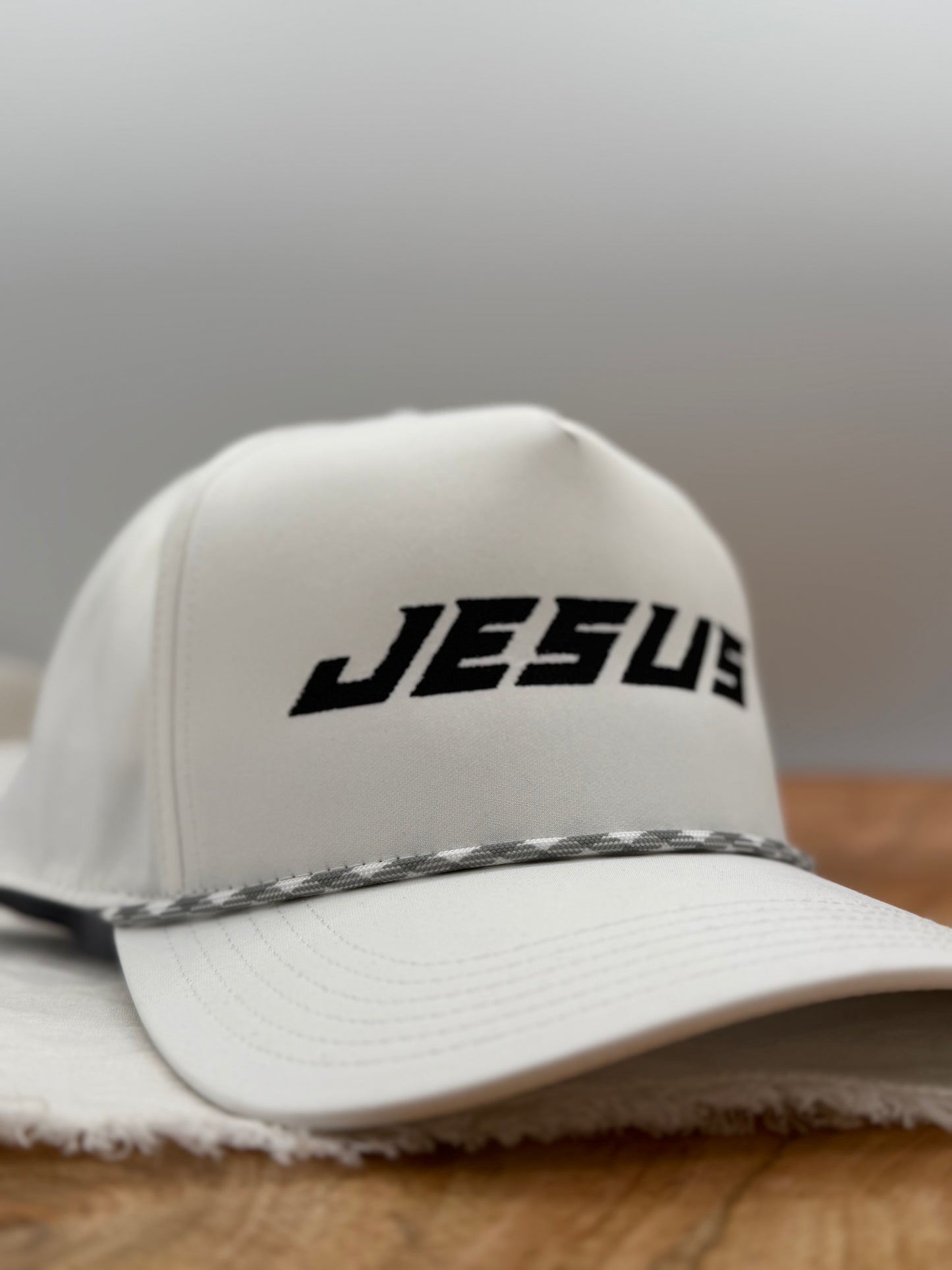 White cap with 'JESUS' embroidered on it in black, featuring a black and white patterned strap, on a wooden surface.
