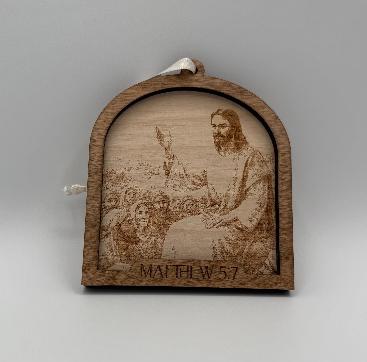 Wooden ornament with a laser engraved scene depicting Jesus praying in front of a crowd. Bible verses Matthew 5:7.