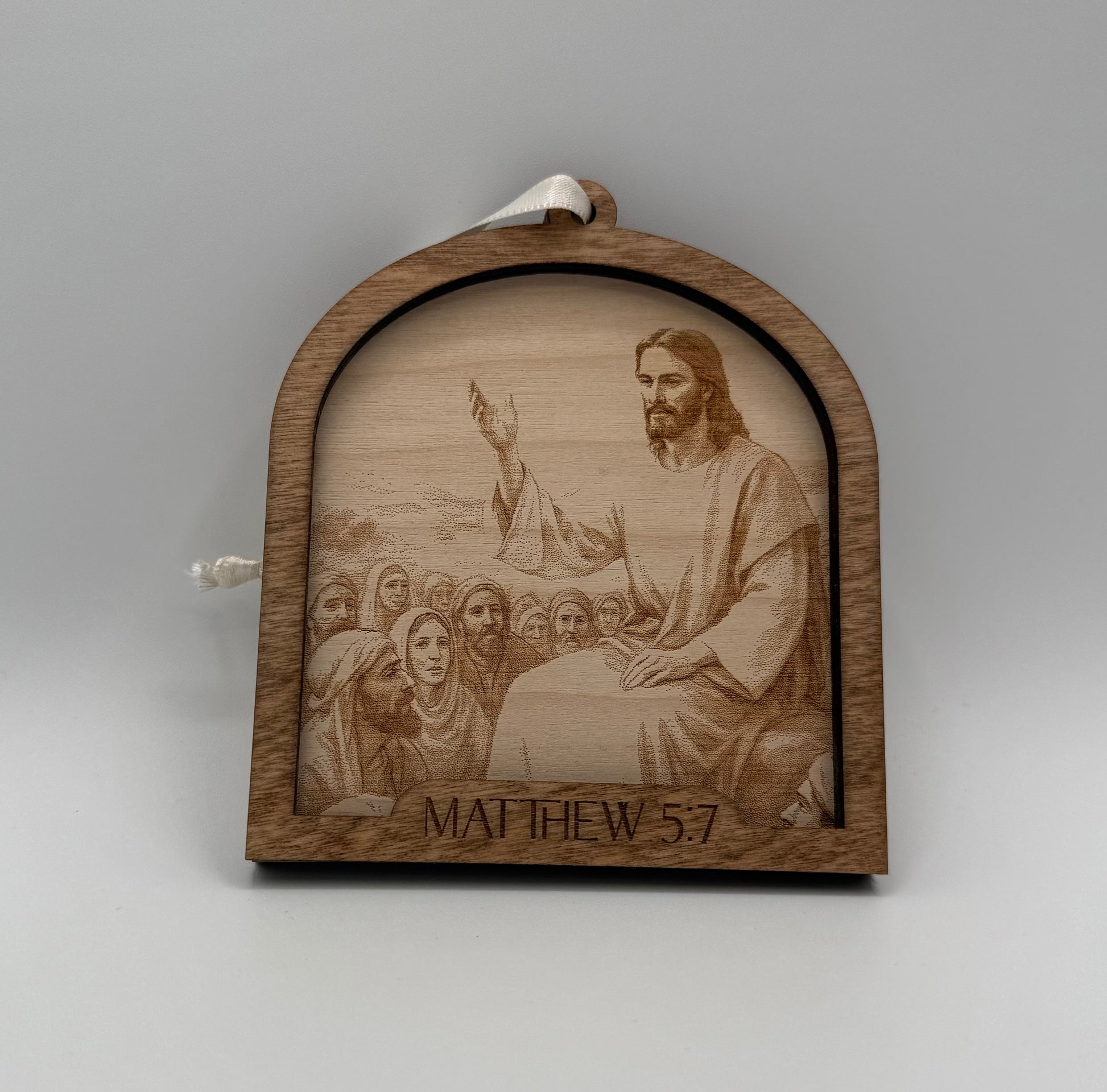Wooden ornament with a laser engraved scene depicting Jesus praying in front of a crowd. Bible verses Matthew 5:7.