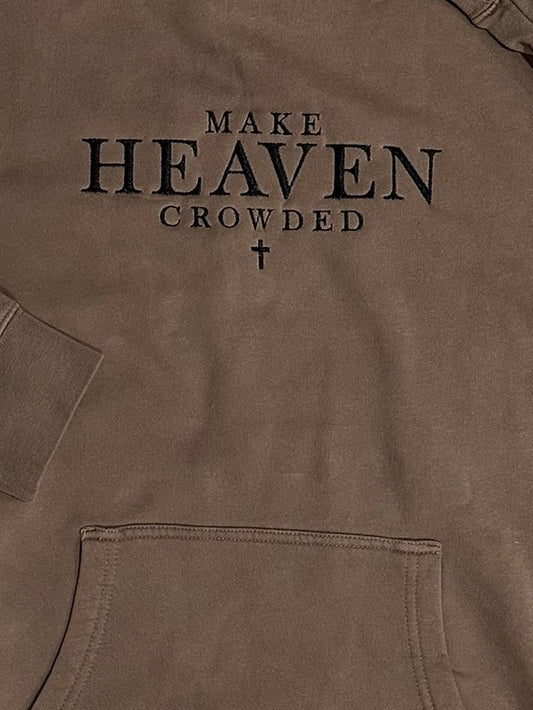 Brown hooded sweatshirt with 'MAKE HEAVEN CROWDED' text and cross symbol embroidered in black on the front center chest. This view is a zoomed in view of the design with hoodie pocket showing.