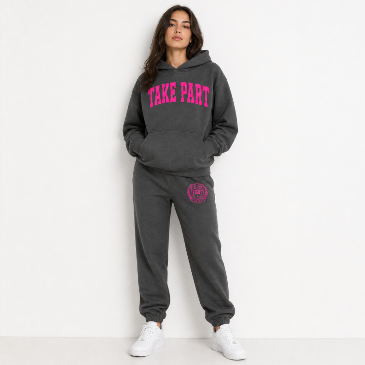 A woman modeling a dark gray two-piece tracksuit with bright pink graphics. The hoodie features the words 'TAKE PART' in pink arched lettering across the chest, and the matching sweatpants have a pink circular graphic on the left thigh.