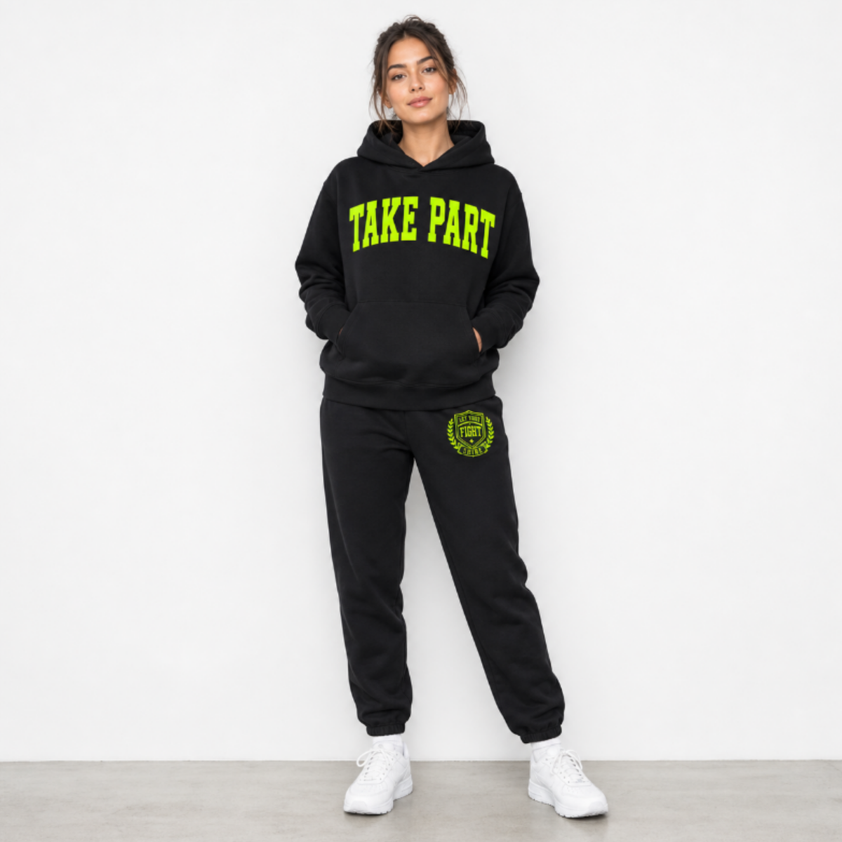 A woman modeling a black sweatshirt and matching sweatpants set. The hoodie features 'TAKE PART' printed in bright neon green across the chest, and the sweatpants have a matching green circular crest graphic on the left thigh.