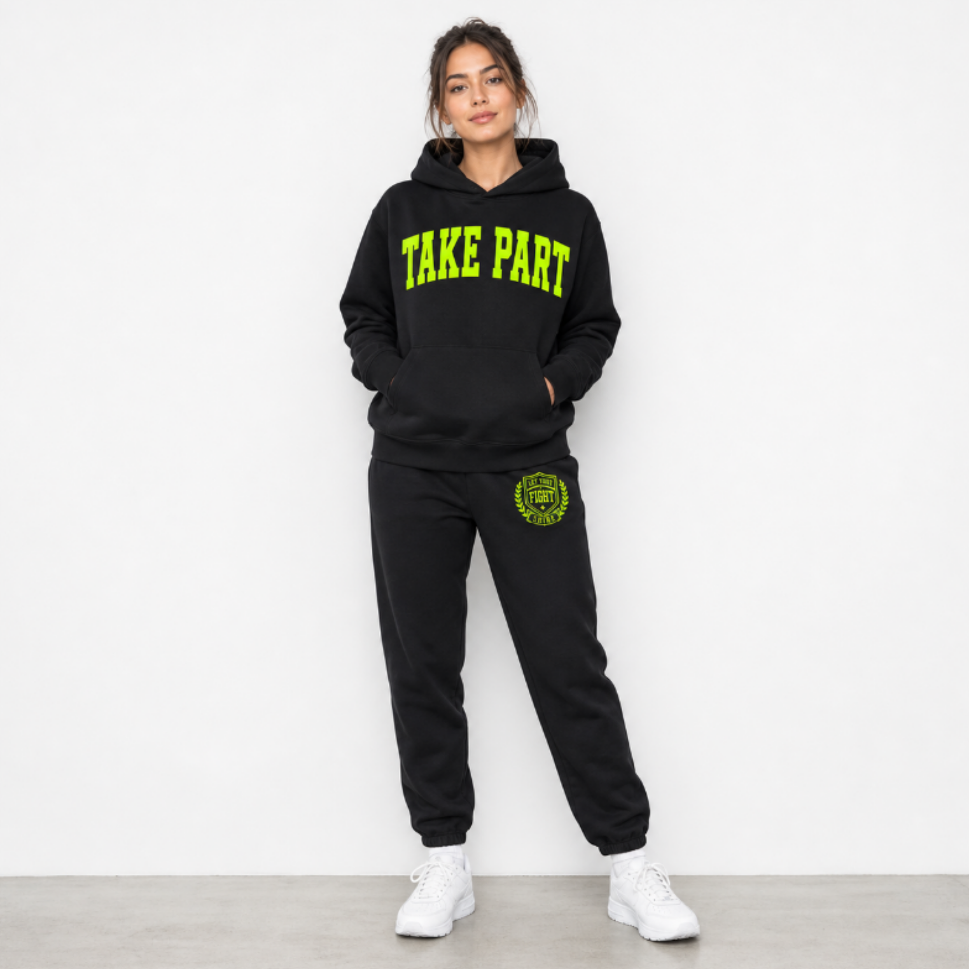 A woman modeling a black sweatshirt and matching sweatpants set. The hoodie features 'TAKE PART' printed in bright neon green across the chest, and the sweatpants have a matching green circular crest graphic on the left thigh.