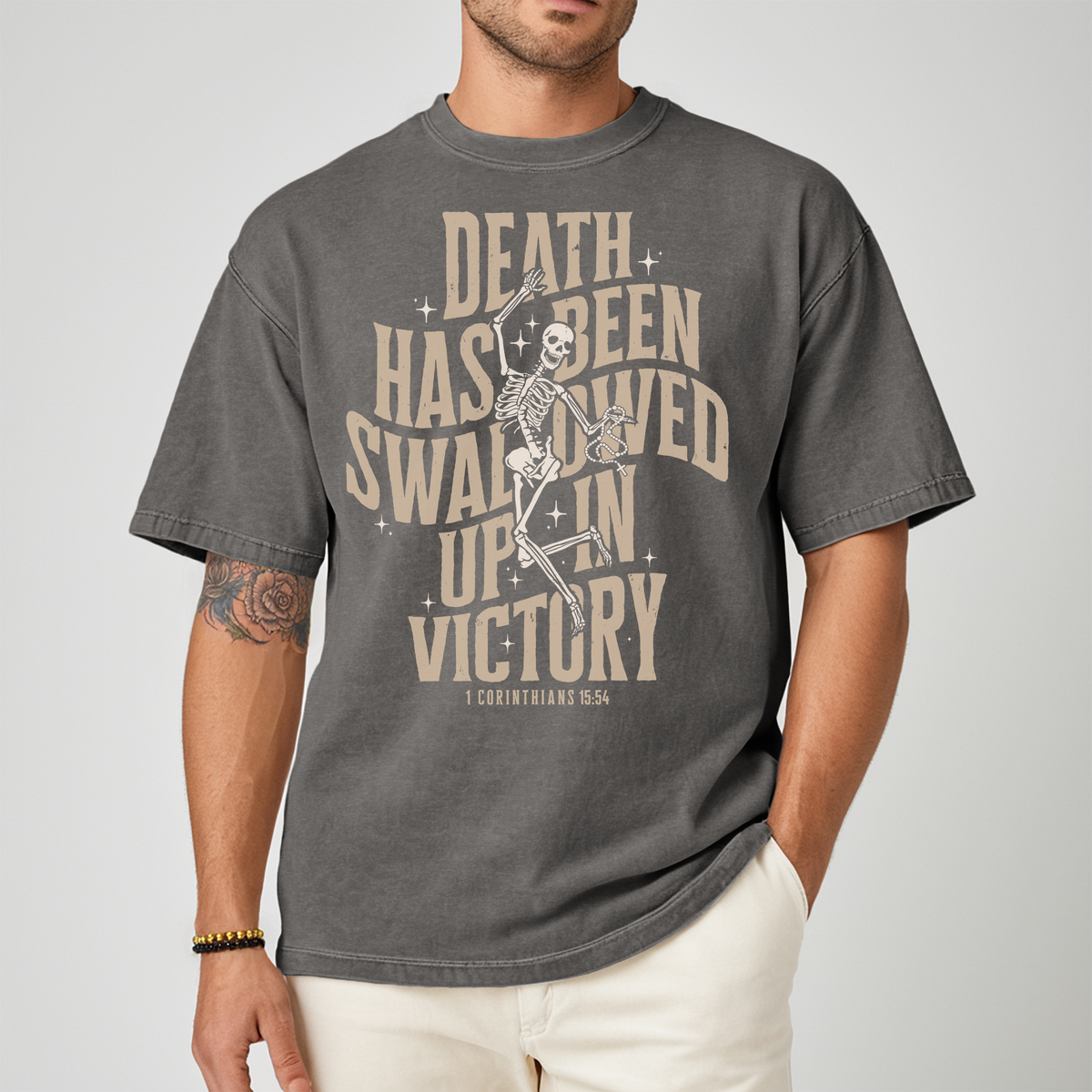 A man wearing a charcoal gray graphic t-shirt with a beige skeleton and text design that reads 'DEATH HAS BEEN SWALLOWED UP IN VICTORY 1 CORINTHIANS 15:54'.