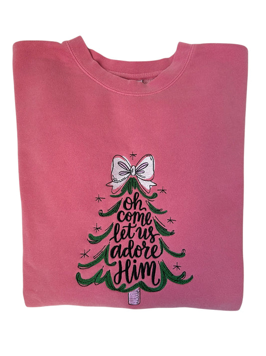 Pink sweatshirt with Christmas tree design and text going through the center of the tree that says 'Oh come let us adore Him' him embroidered on a white background. Sweatshirt is folded front view.