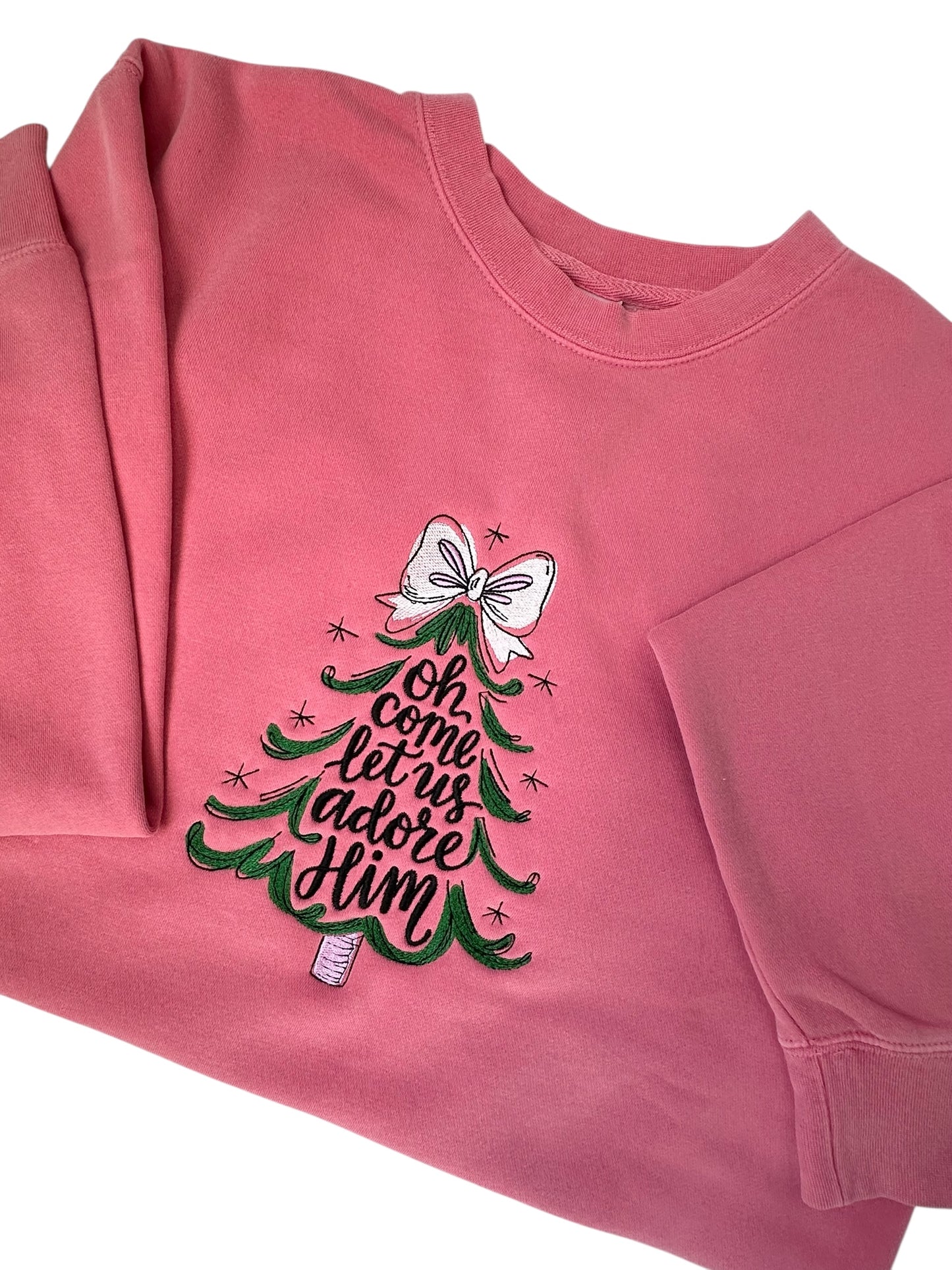 Pink sweatshirt with Christmas tree design and text going through the center of the tree that says 'Oh come let us adore Him' him embroidered front center chest. The tree features a white bow, the tree is green, and the words are black. Sweatshirt is folded in half and sleeves are arranged.