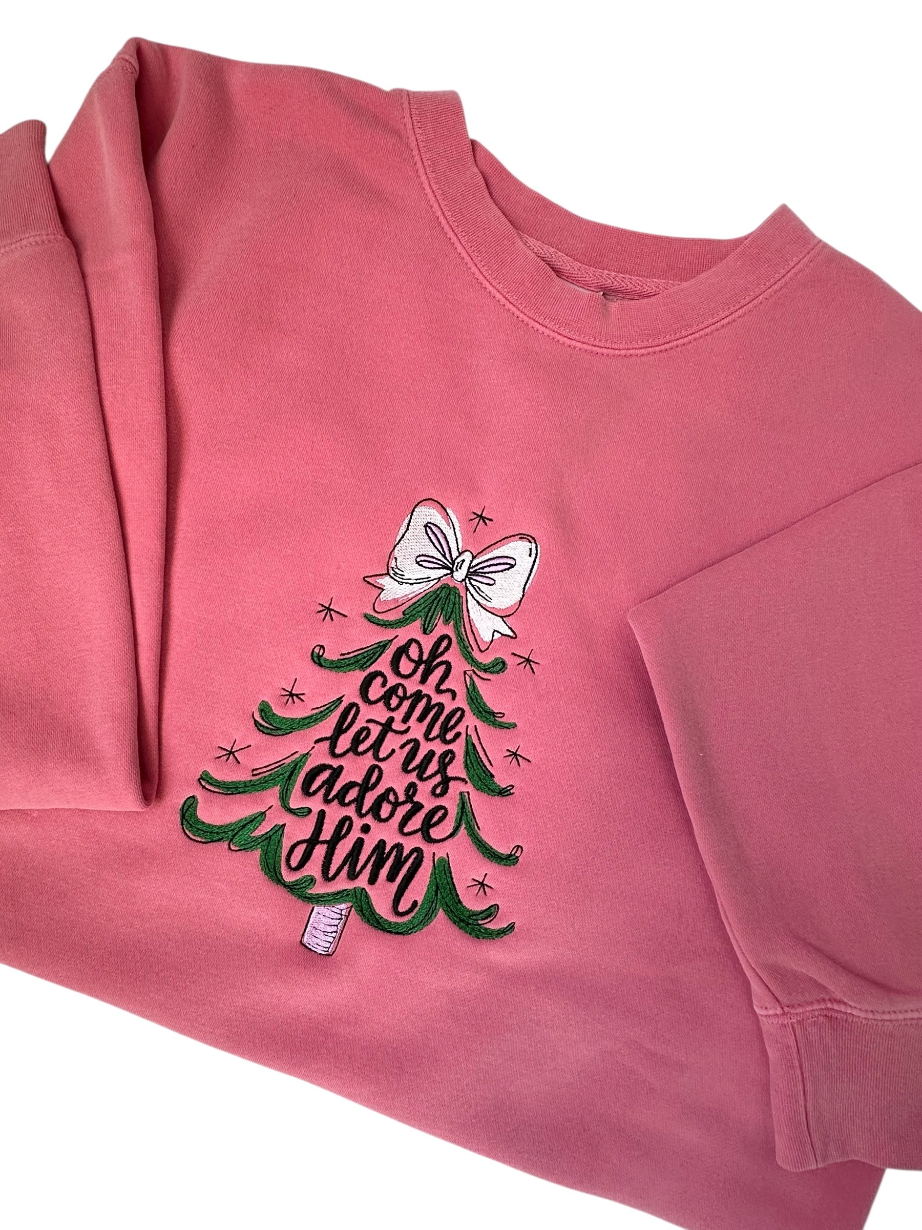 Pink sweatshirt with Christmas tree design and text going through the center of the tree that says 'Oh come let us adore Him' him embroidered front center chest. The tree features a white bow, the tree is green, and the words are black. Sweatshirt is folded in half and sleeves are arranged.