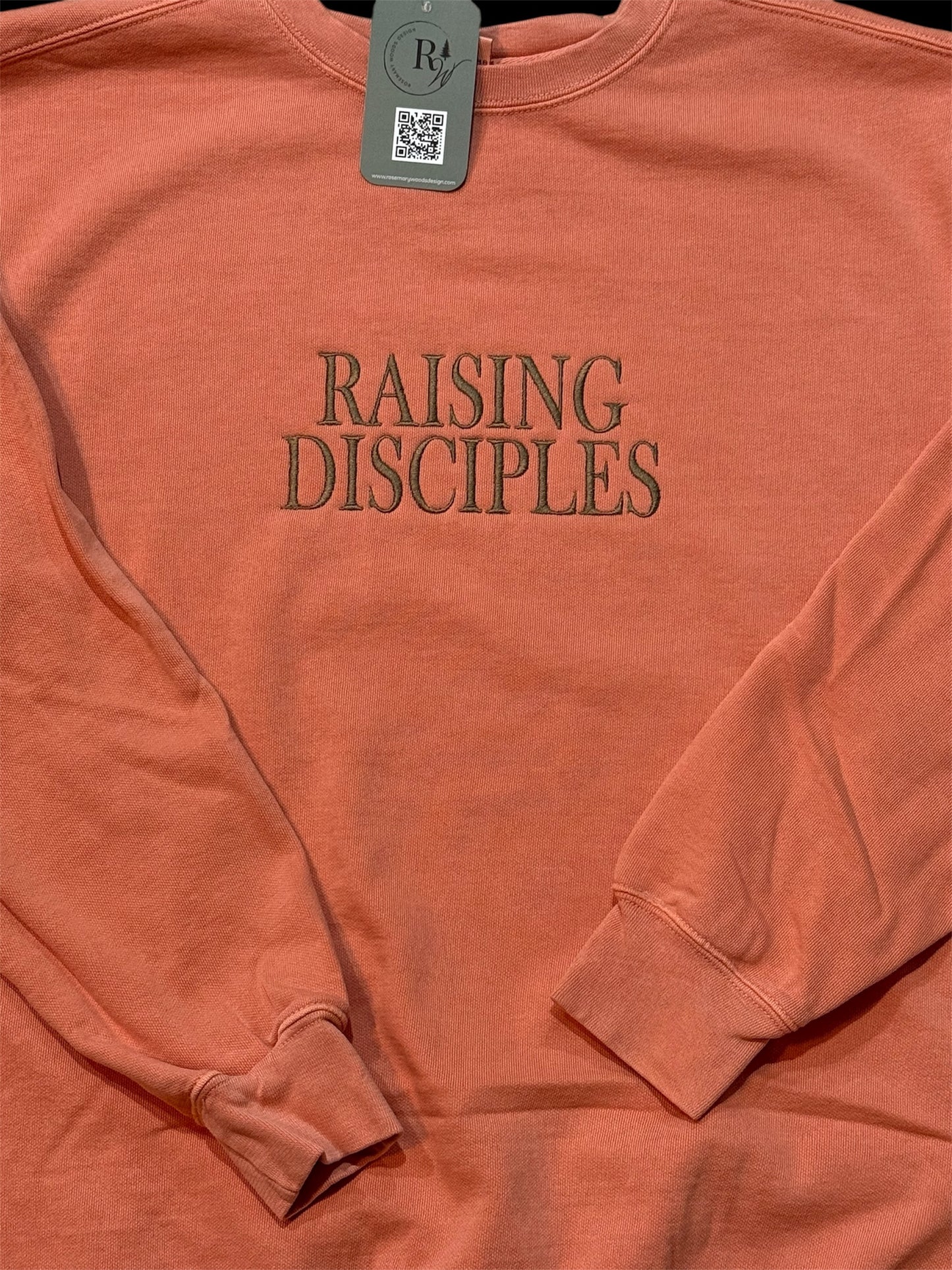 Coral sweatshirt with 'RAISING DISCIPLES' text embroidered in capital letters on the front center chest in brown. Zoomed in view with folded sleeves.