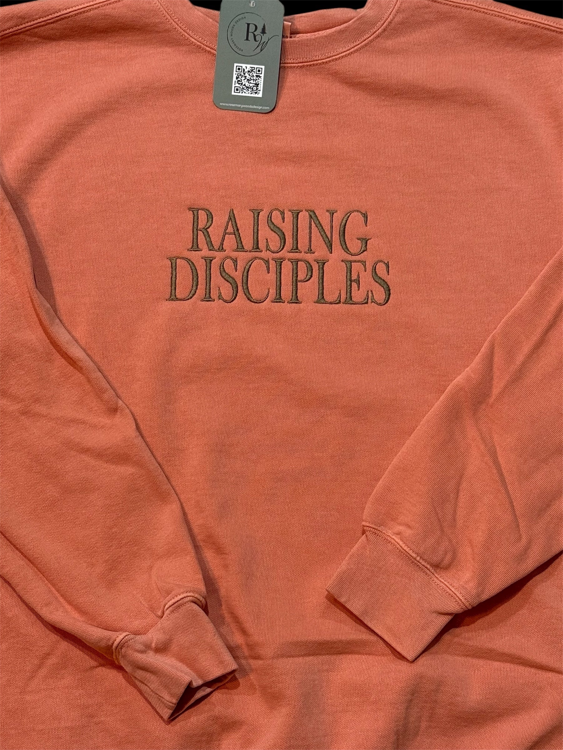 Coral sweatshirt with 'RAISING DISCIPLES' text embroidered in capital letters on the front center chest in brown. Zoomed in view with folded sleeves.