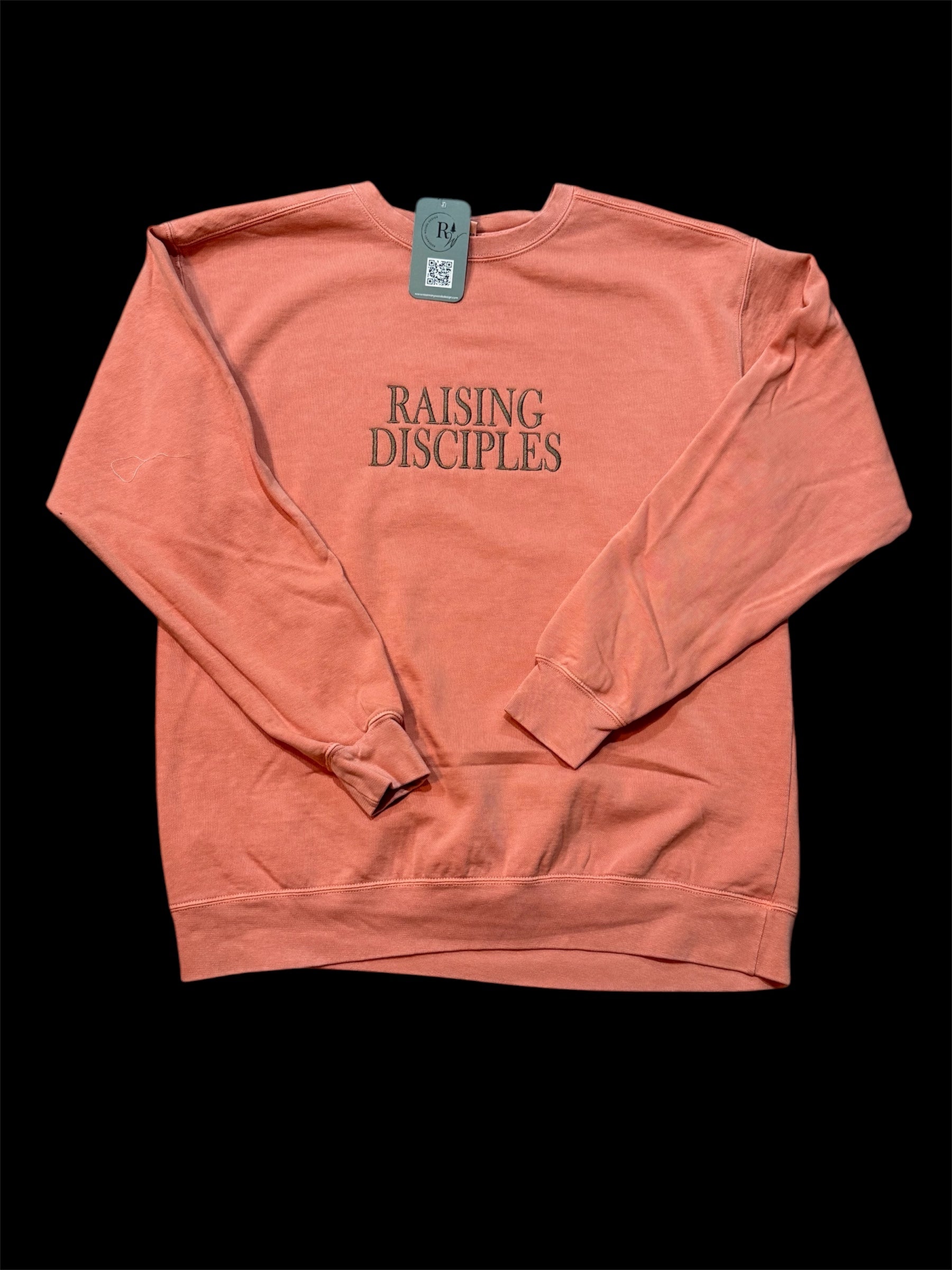 Coral sweatshirt with the text 'Raising Disciples' embroidered in capital letters in brown. This view is zoomed out with the entire sweatshirt showing with sleeves folded on top.