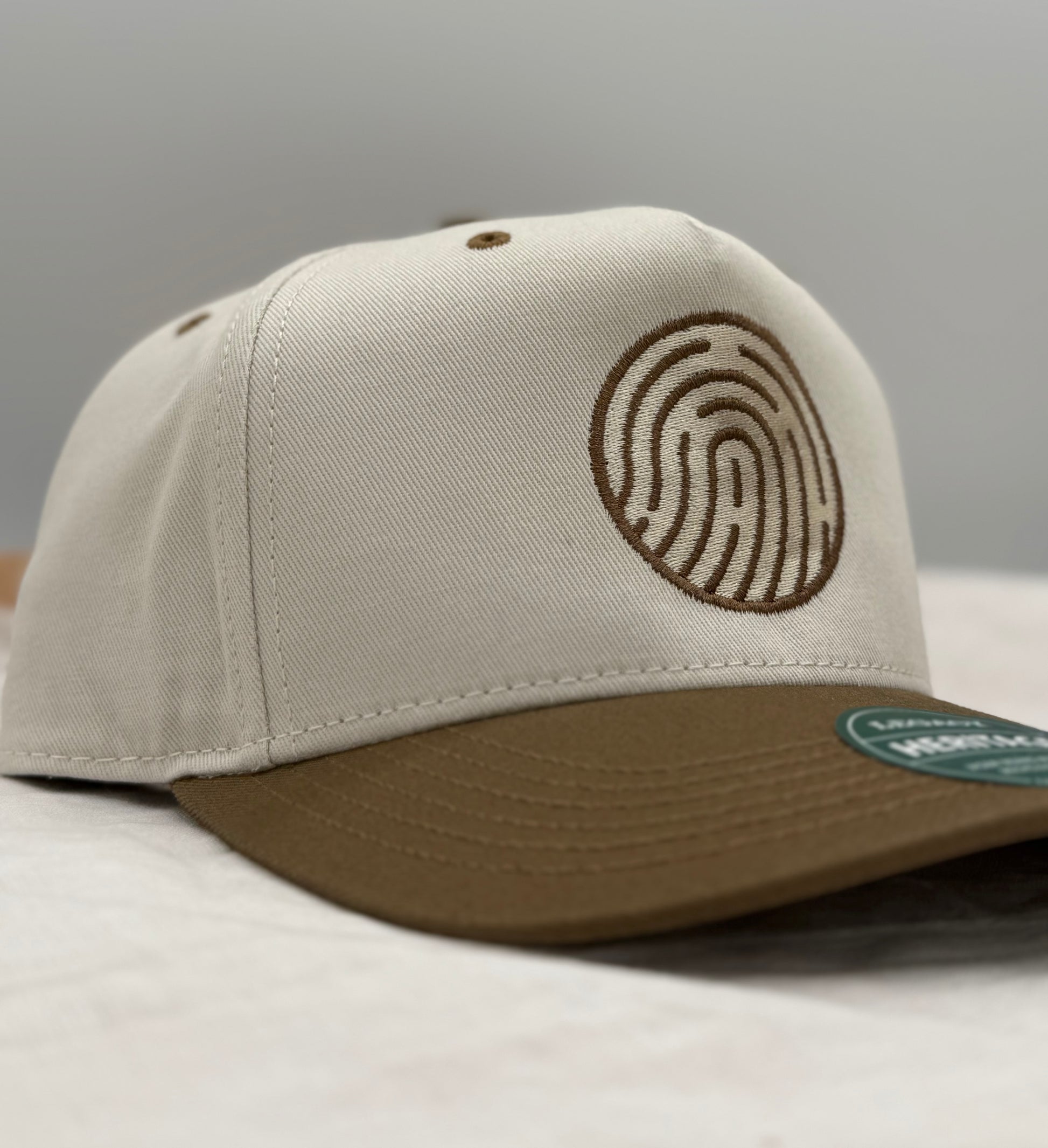 Beige and brown 2 tone hat side profile with a Soma church fingerprint logo embroidered in brown on a gray background