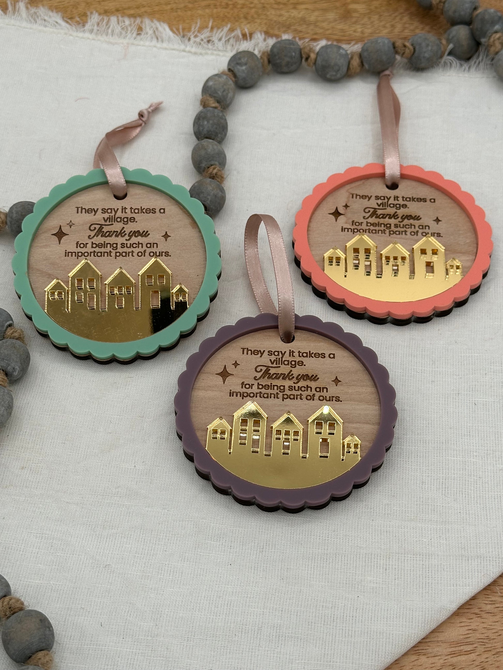 Three circle wood ornament with text laser engraved that says, 'They say it takes a village thank you for being such an important part of ours.' The ornaments feature an acrylic scalloped outline and a golden mirrored village.