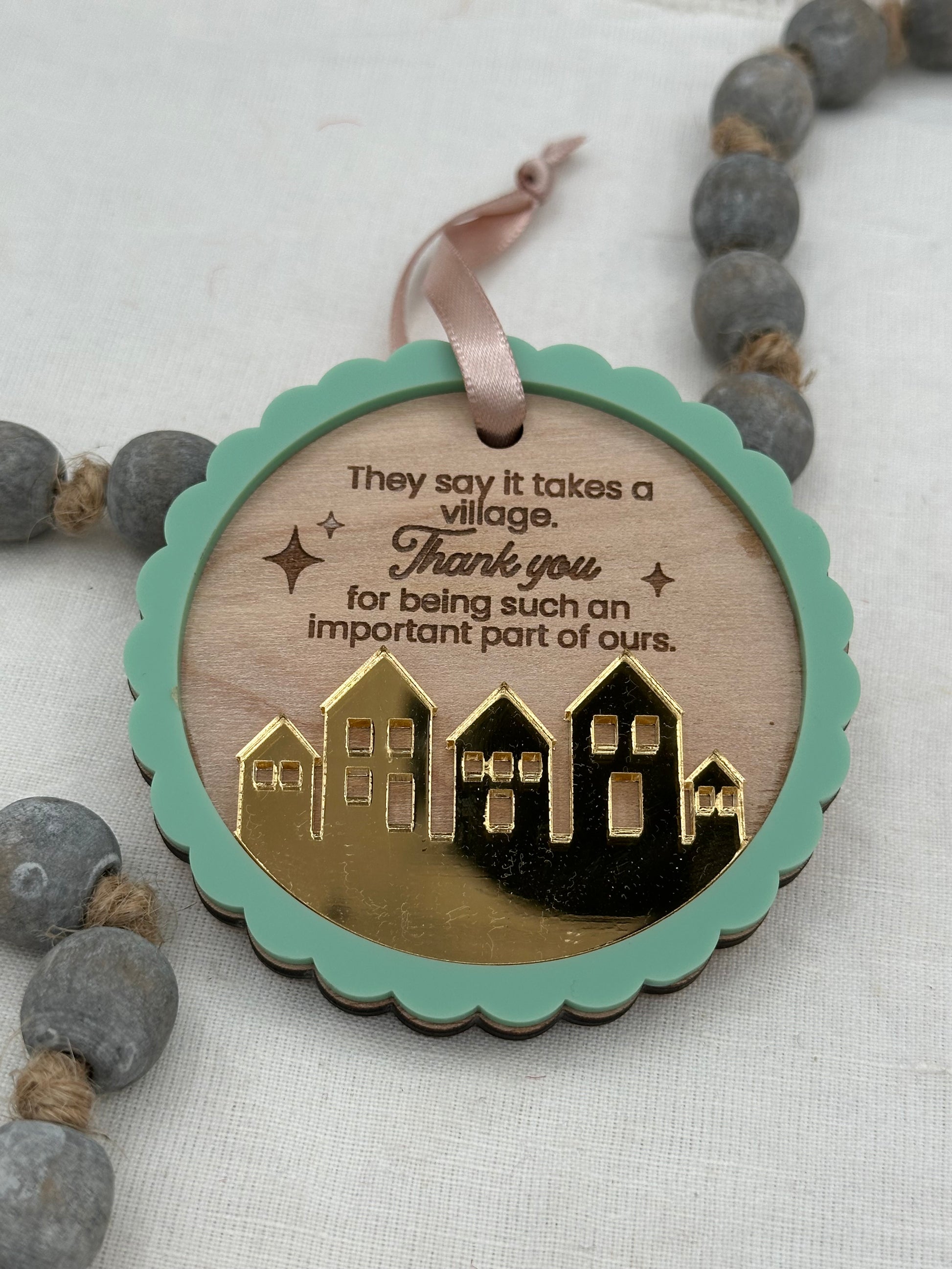 Circle wood ornament with text laser engraved that says 'They say it takes a village thank you for being such an important part of ours.' The ornament features an acrylic green scalloped outline and a golden mirrored village.