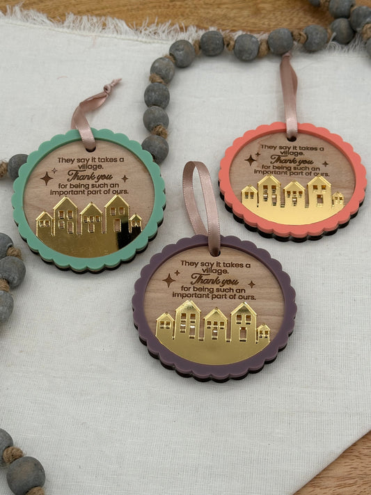 Three decorative circle ornaments with text laser engraved that says 'They say it takes a village thank you for being such an important part of ours.' The ornament features an acrylic scalloped outline and a golden mirrored village.