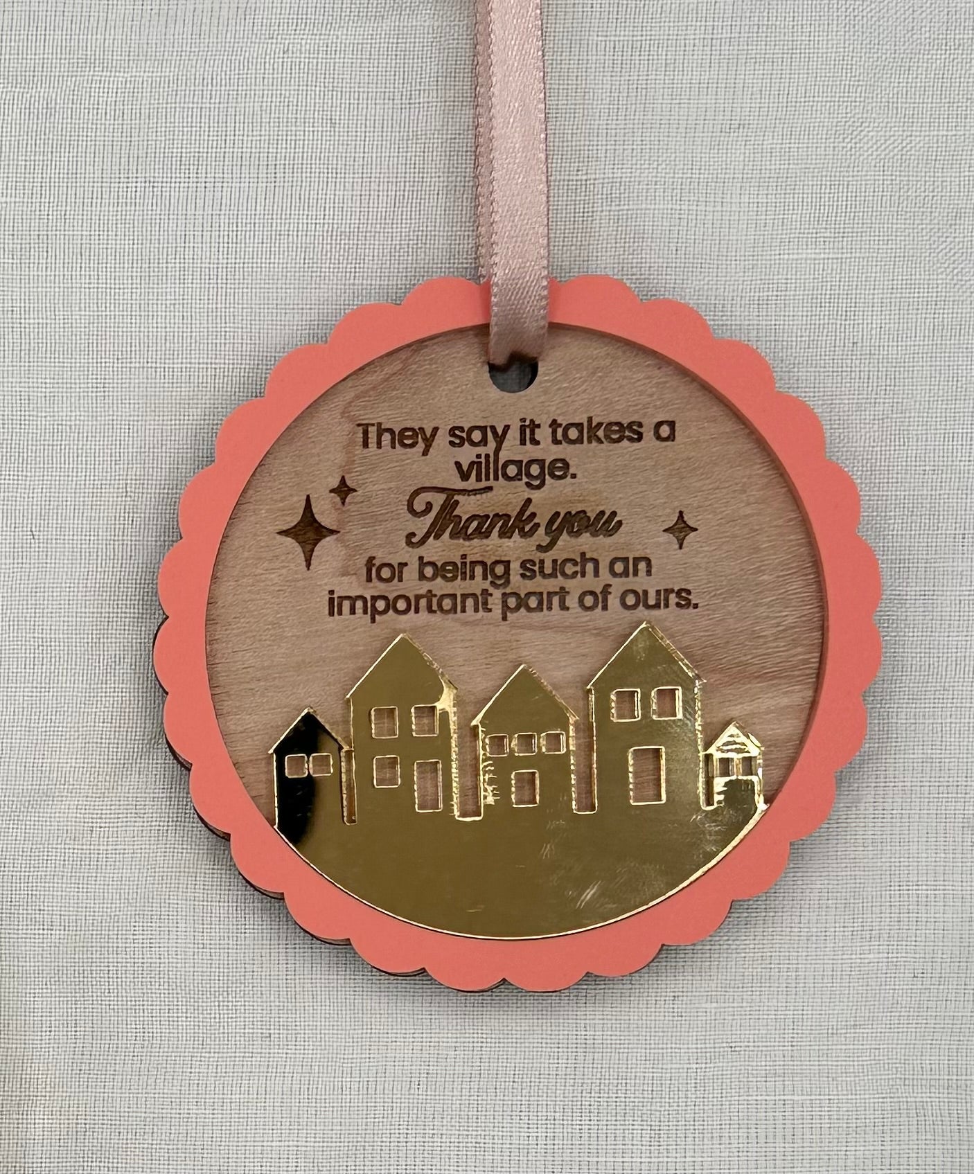 Circle wood ornament with text laser engraved that says 'They say it takes a village thank you for being such an important part of ours.' The ornament features an acrylic pink scalloped outline and a golden mirrored village.