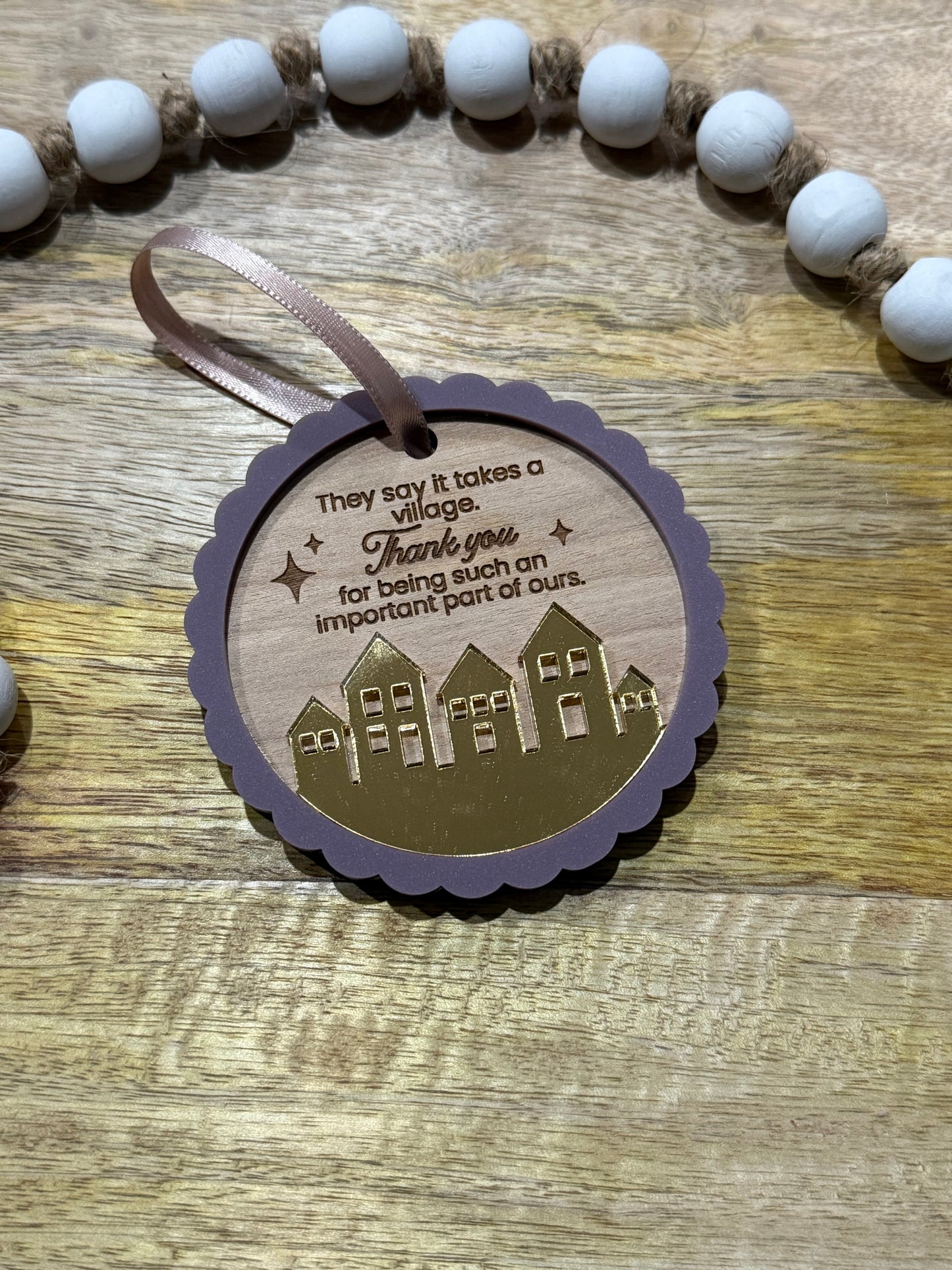 Circle wood ornament with text laser engraved that says 'They say it takes a village thank you for being such an important part of ours.' The ornament features an acrylic purple scalloped outline and a golden mirrored village.