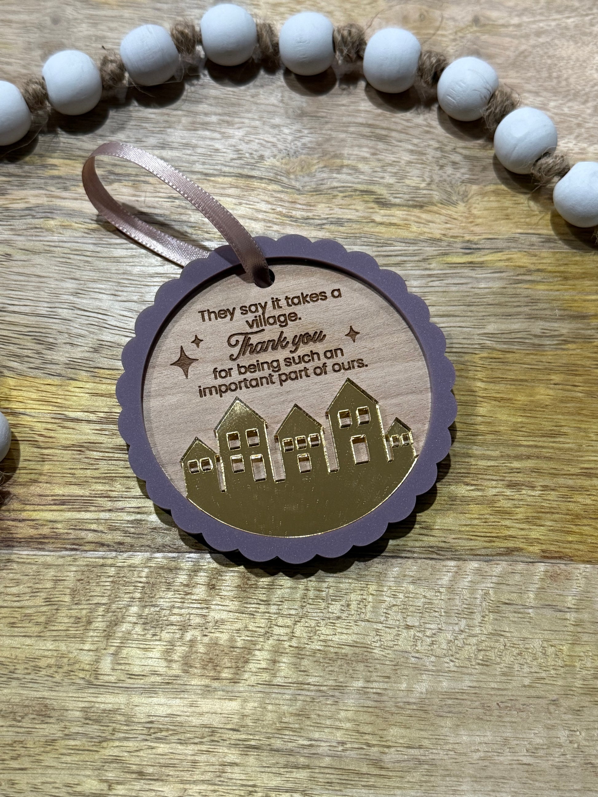 Circle wood ornament with text laser engraved that says 'They say it takes a village thank you for being such an important part of ours.' The ornament features an acrylic purple scalloped outline and a golden mirrored village.